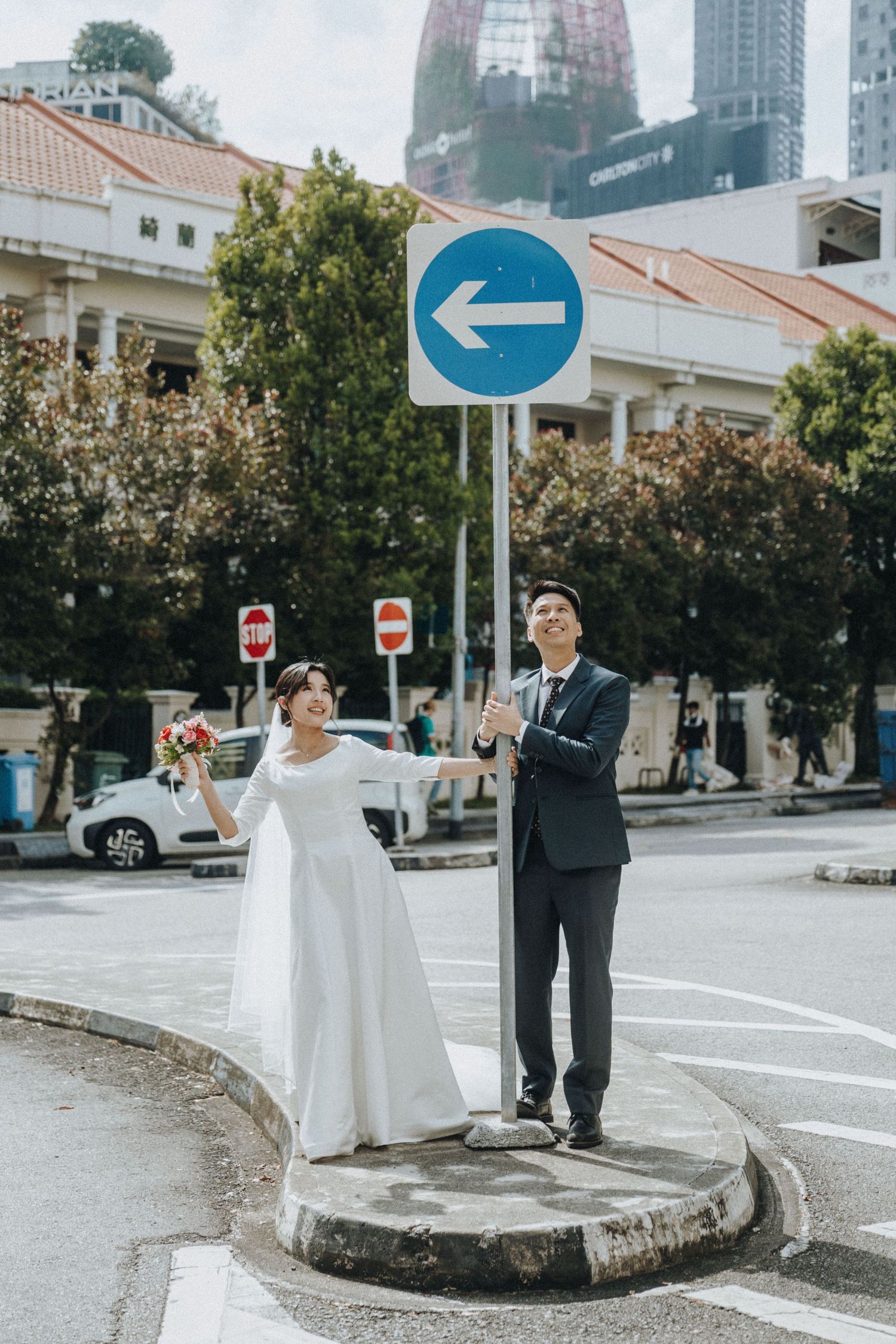 Bride and groom pose on a traffic island beside a blue arrow sign in an urban Singapore street setting.