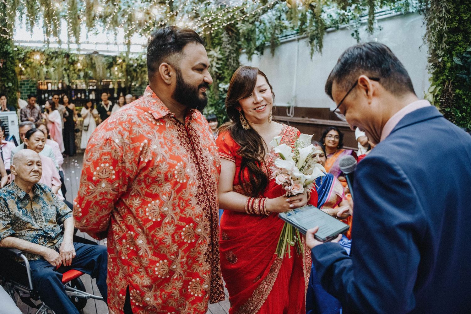 Bride in red saree and groom in floral shirt greet guest during wedding ceremony.