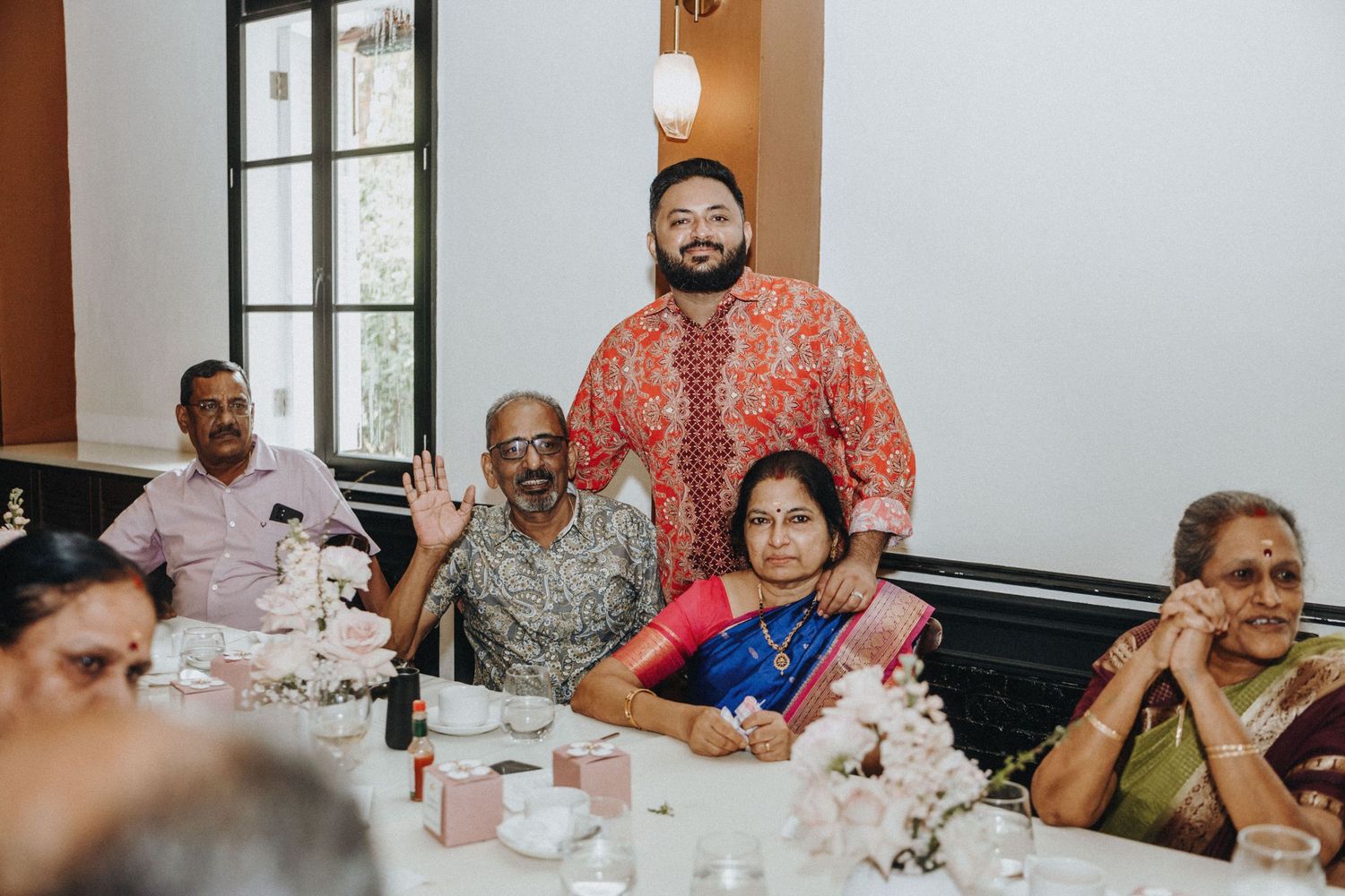Groom in red Indian attire poses with seated family members at a wedding reception dinner table.