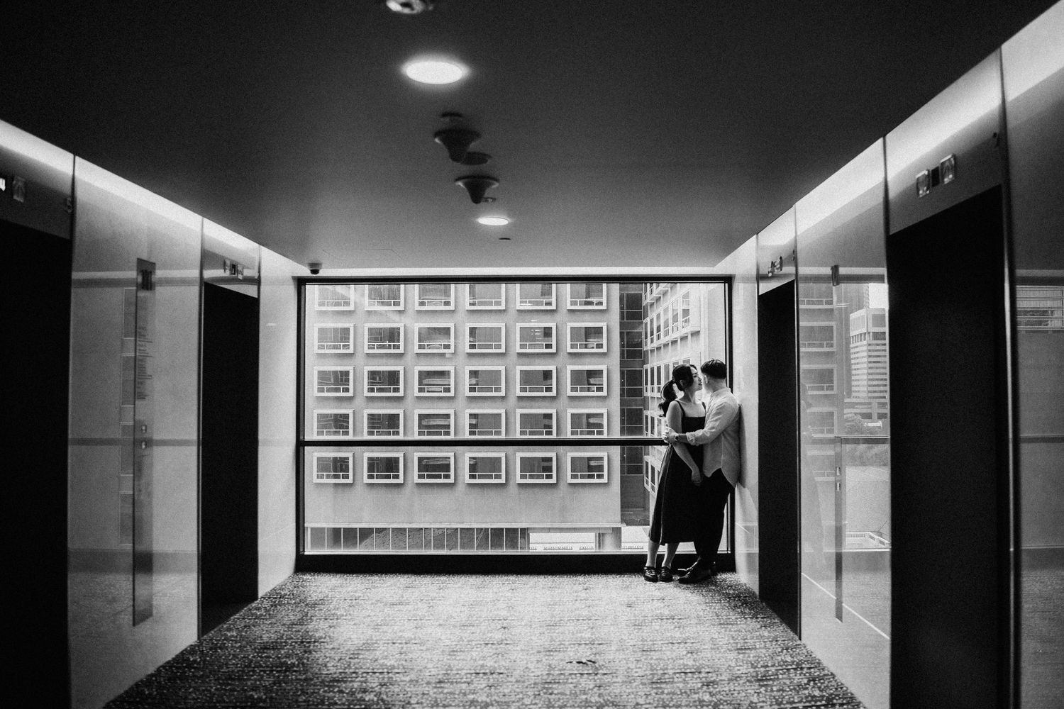 Black and white photo of a couple embracing in a modern building corridor with elevator doors and a grid-patterned wall.