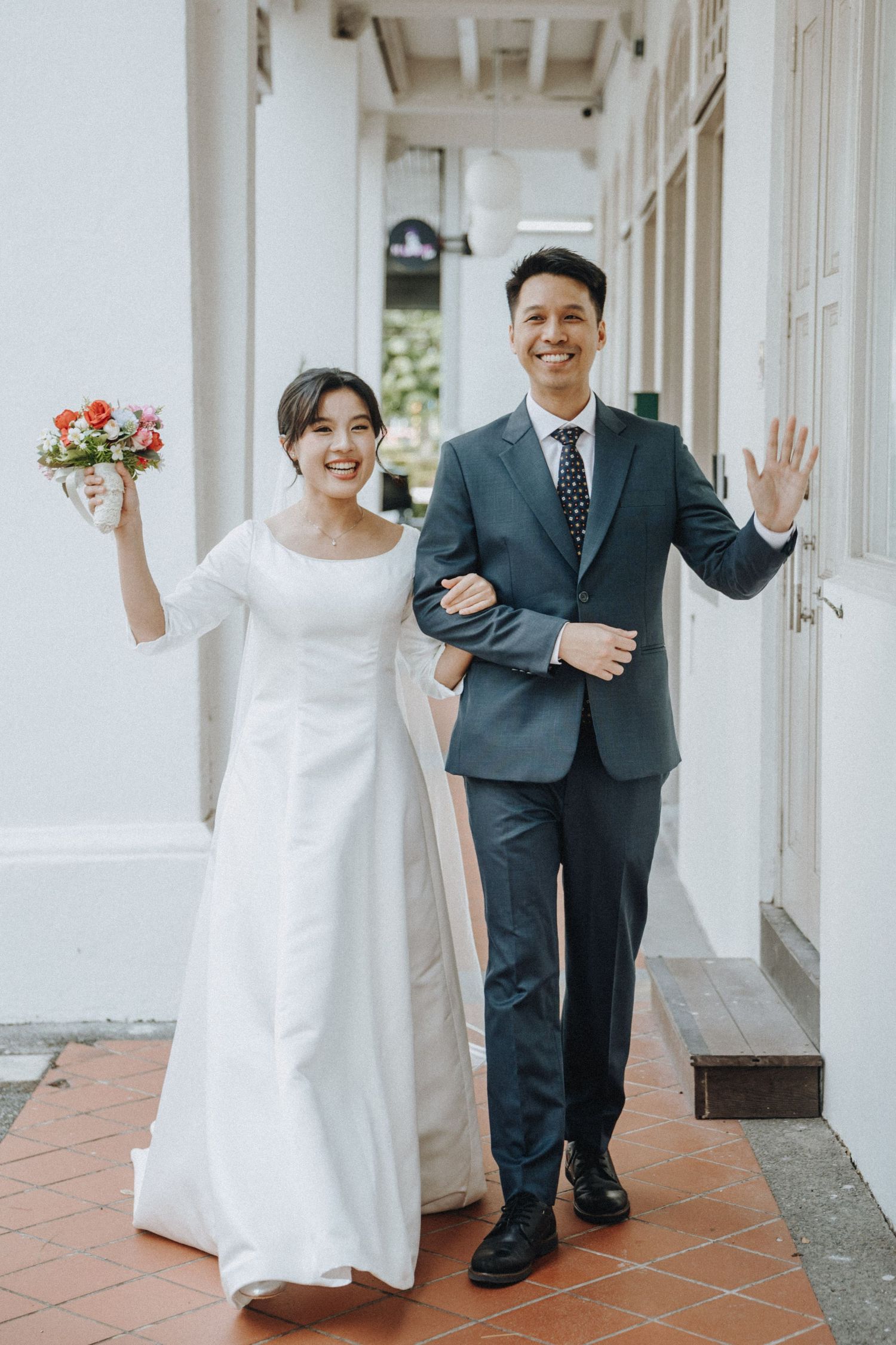 Happy bride and groom walking arm in arm, waving, bride holding bouquet.
