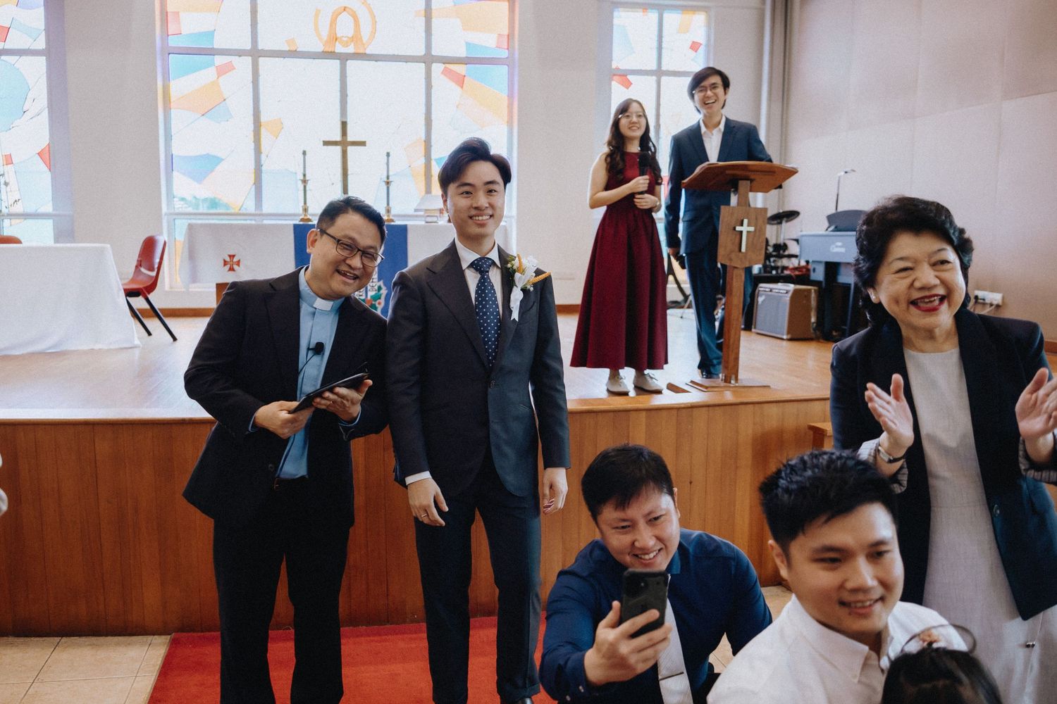 Groom poses and smiles with wedding guests inside a bright church during a wedding celebration.