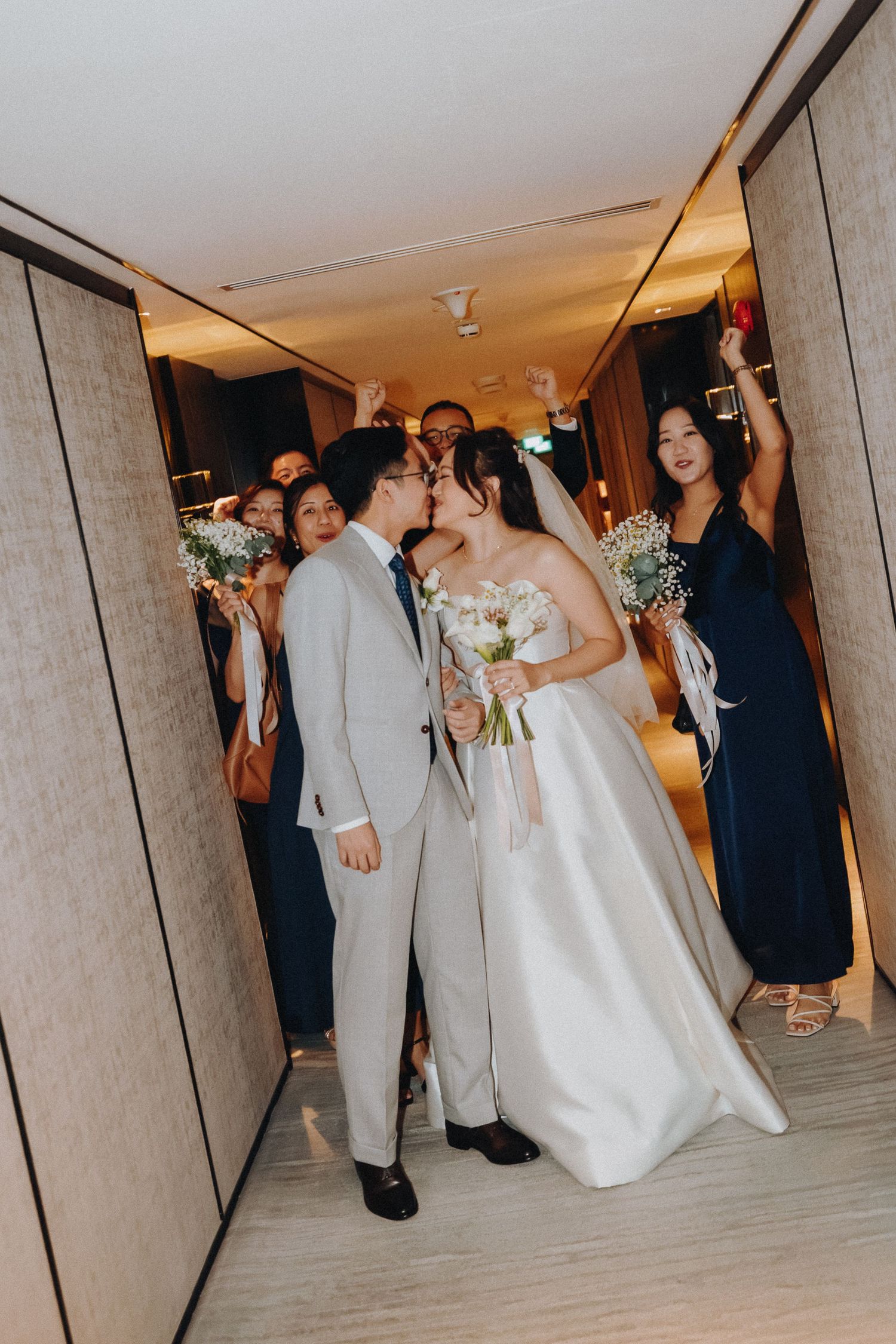 Wedding party walks down a modern hotel hallway with the couple in white and gray attire and attendants in navy dresses.