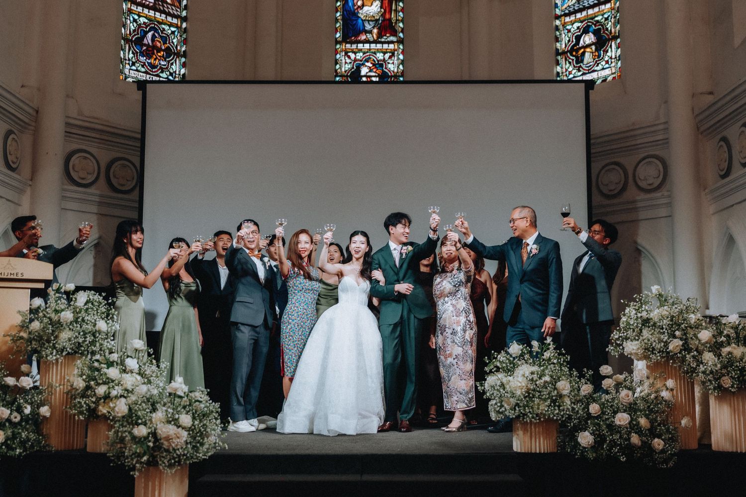 Bride and groom celebrate with family and friends on church altar stage surrounded by white floral arrangements.