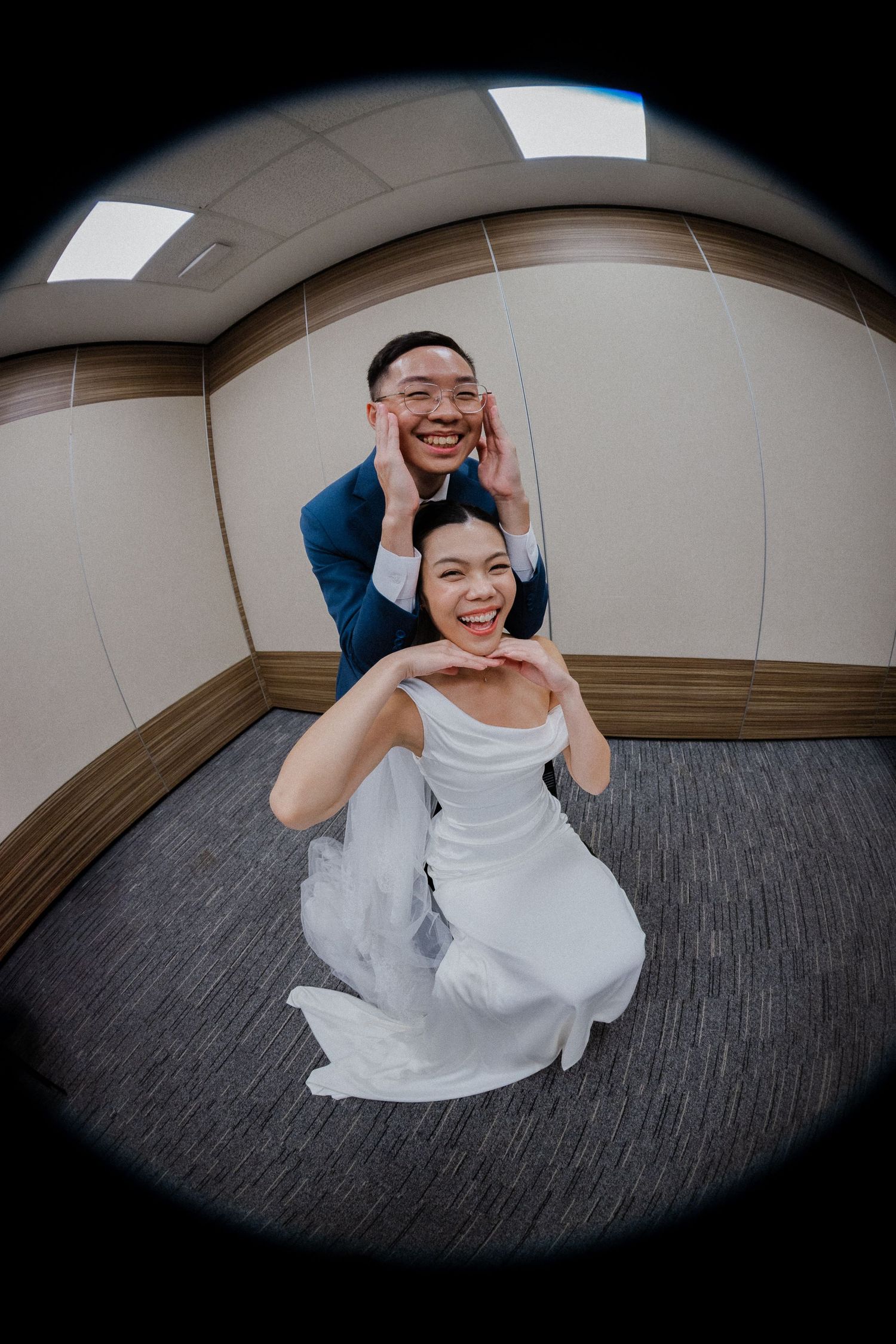 Joyful Asian bride and groom posing playfully in elevator, bride in white gown, groom in blue suit, fisheye lens photo.