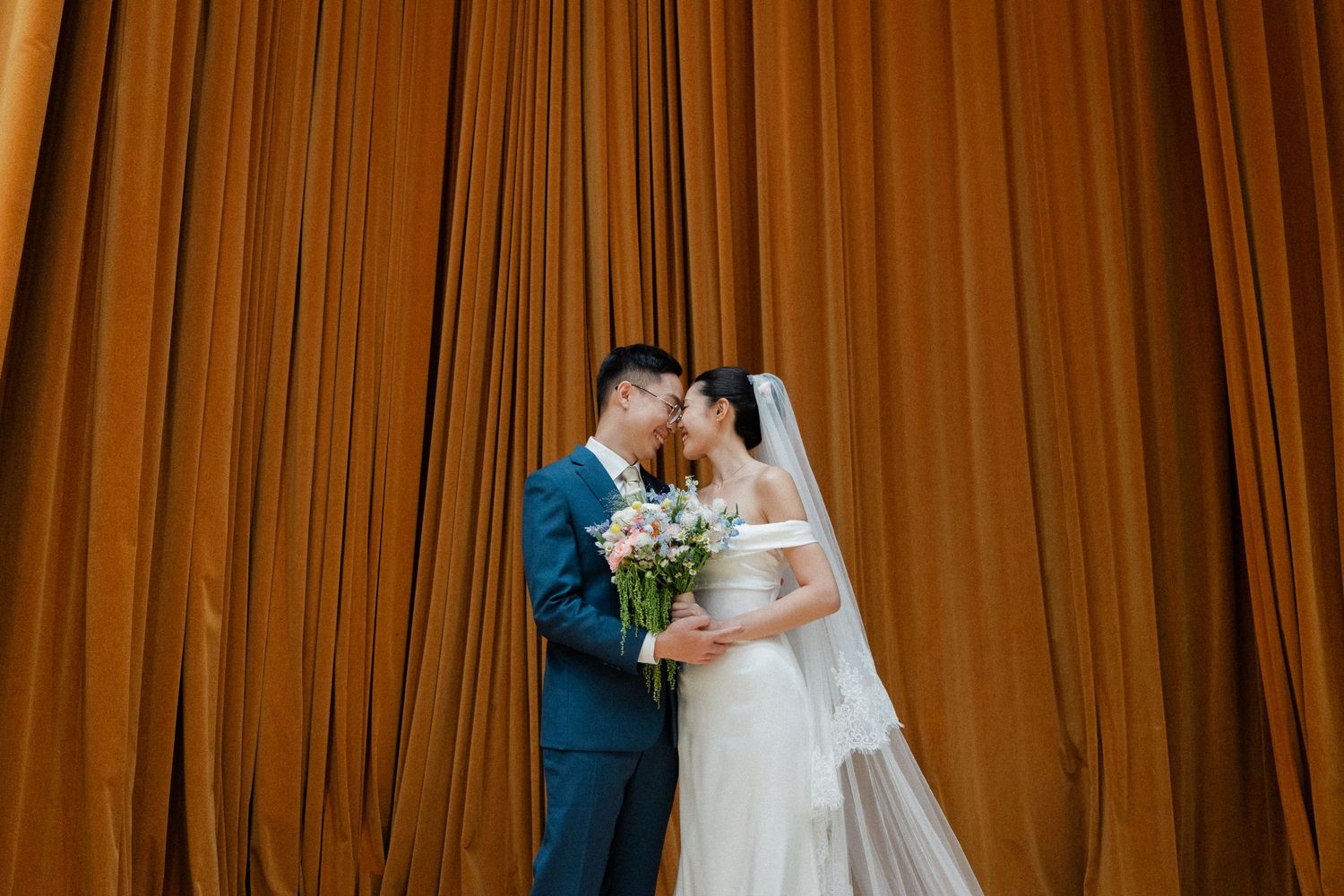 Bride and groom share a romantic kiss in front of golden amber curtains, bride holding a pastel bouquet.