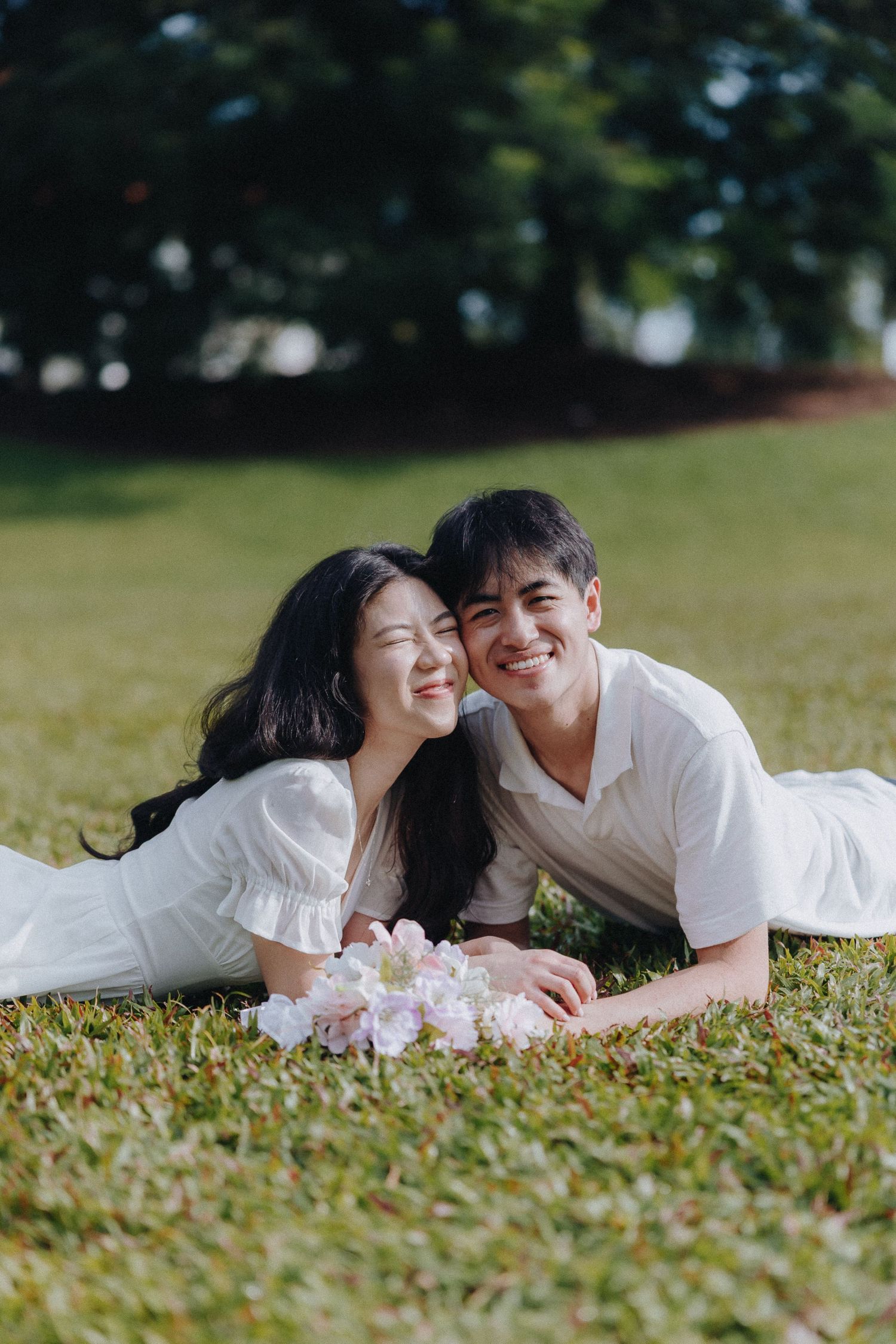 A couple lies on grass sharing laughter with pink flowers between them.