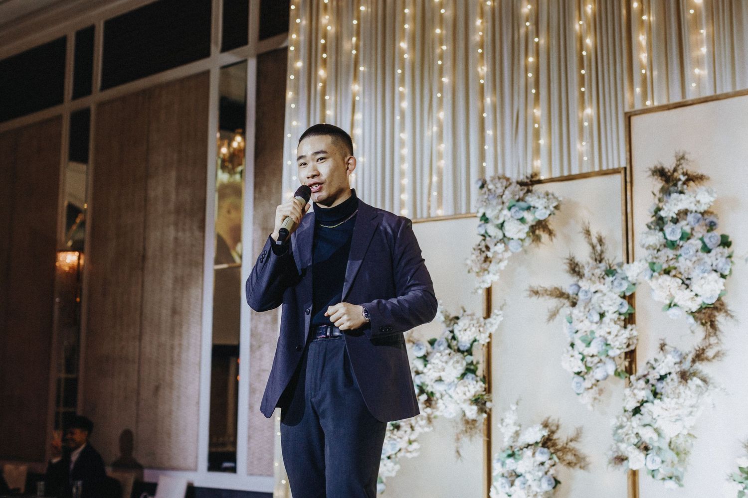 Speaker holds microphone at elegant venue with floral backdrop and string lights.