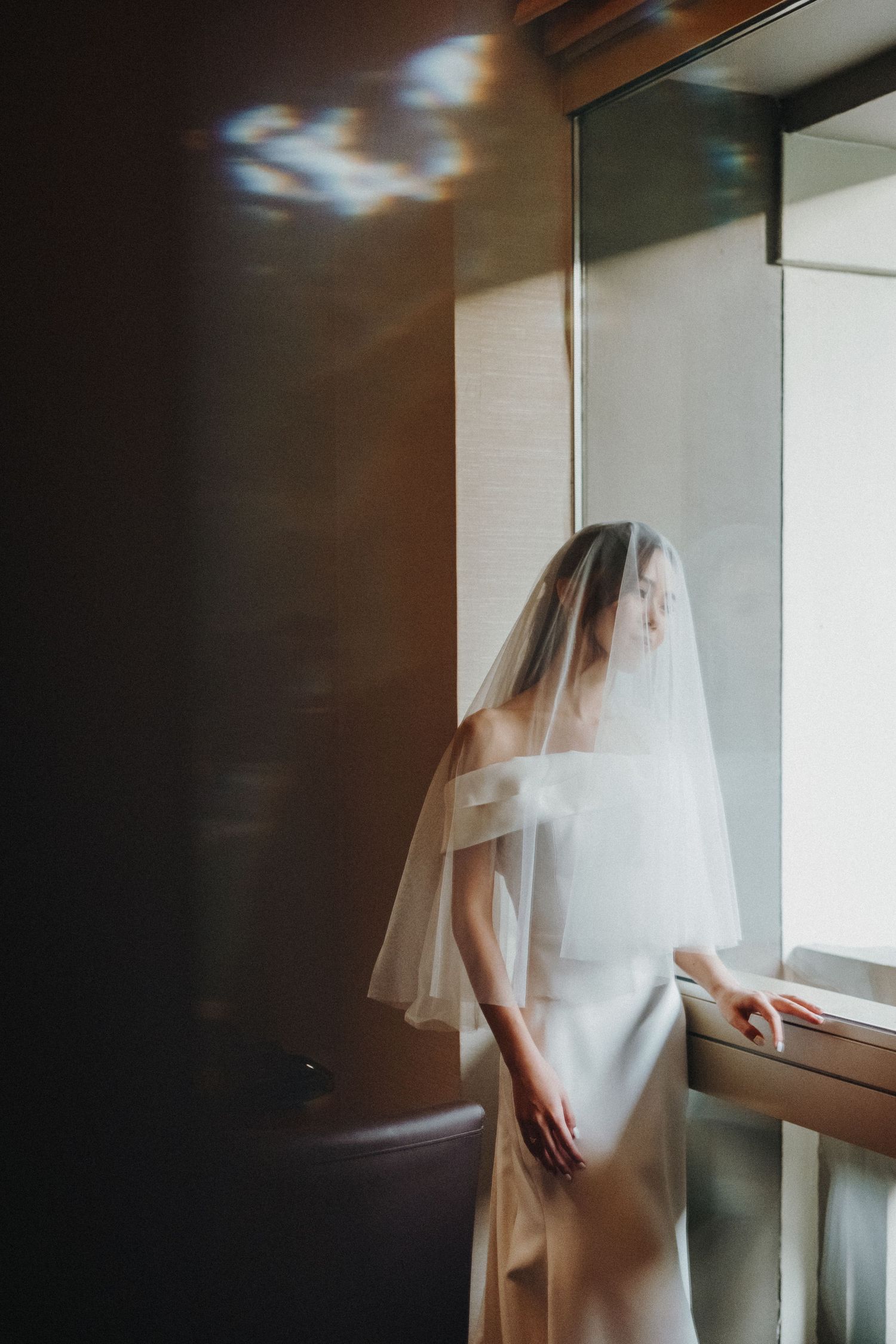 A bride in a flowing veil stands pensively by a window in soft, moody lighting.