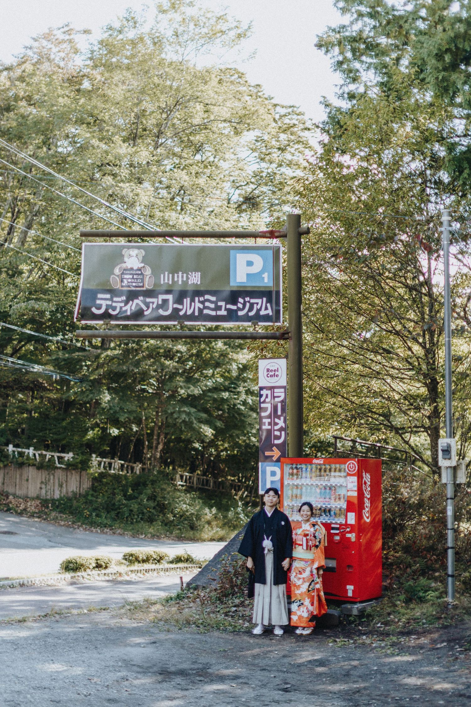 A row of vending machines sits beneath trees on a park pathway with signage in Japanese text.