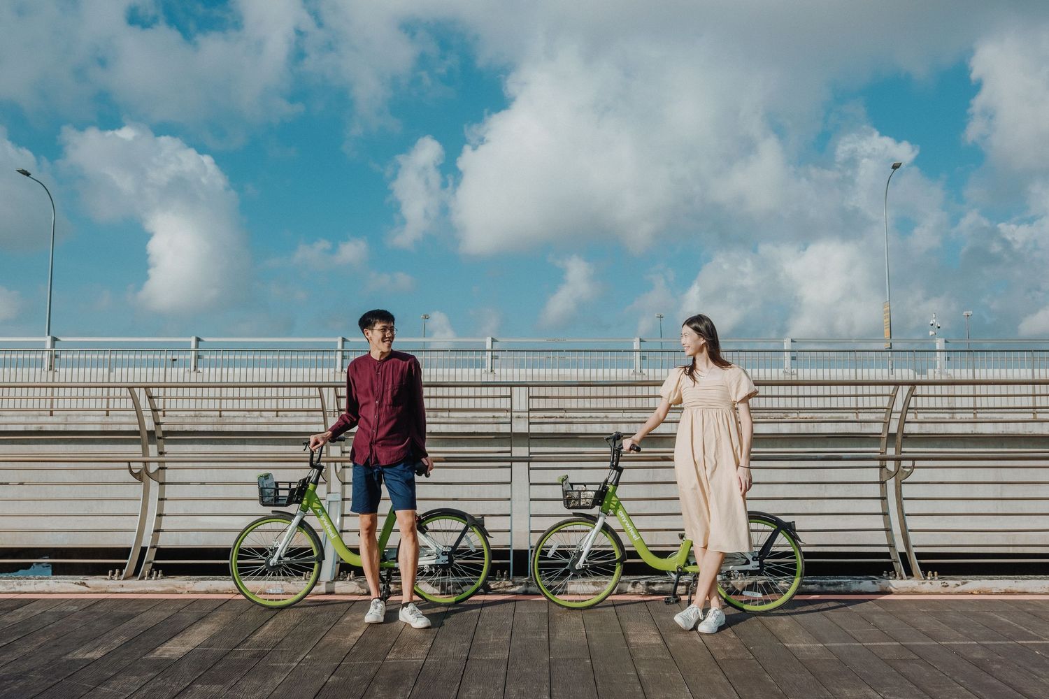 A couple stands with shared bikes on a boardwalk against a cloudy blue sky.