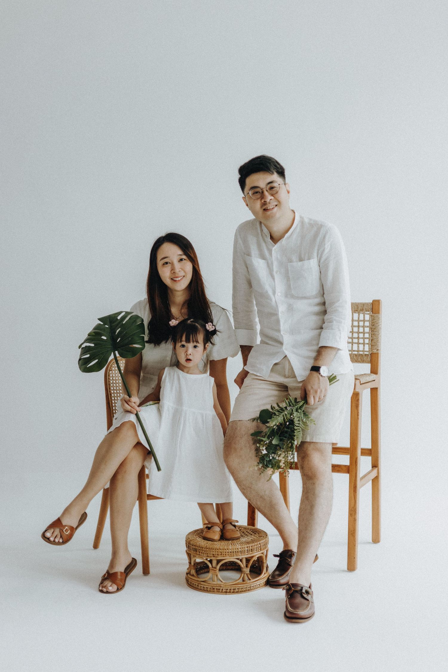 Family in coordinated white outfits poses with plants against light background for minimalist portrait.