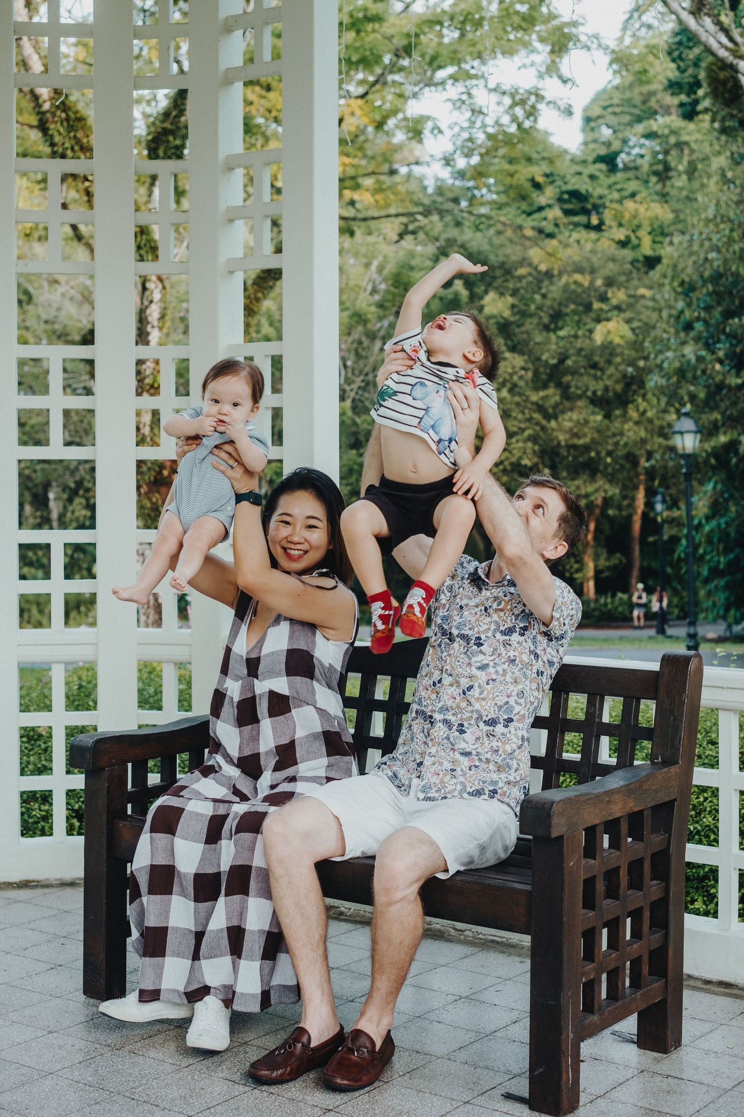 A cheerful family plays together and lifts children while sitting on a wooden bench in a white gazebo.