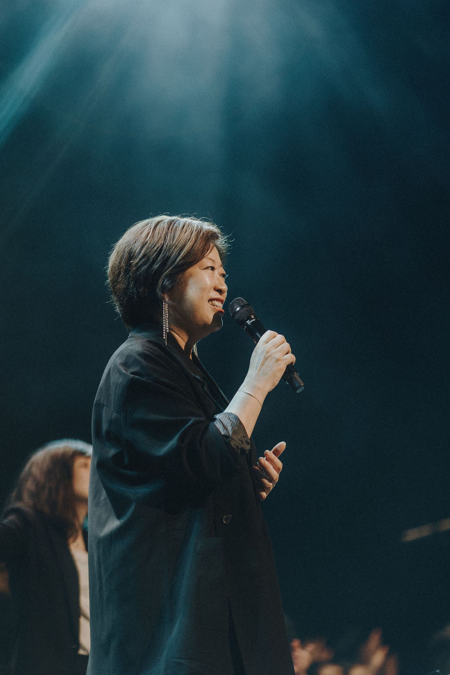Singer performing under dramatic teal stage lighting with emotional expression and dark backdrop.