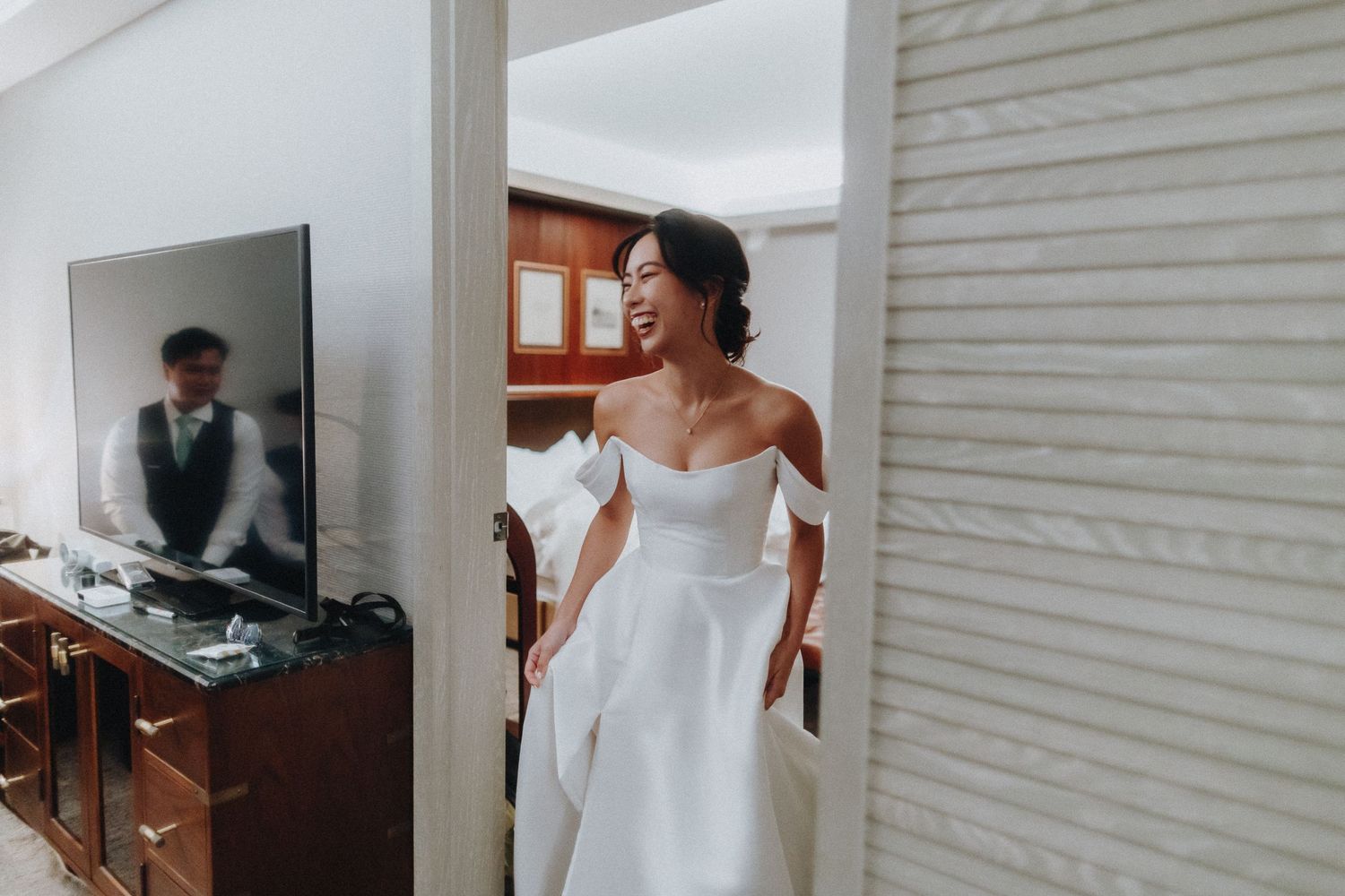 Elegant bride in a modern off-shoulder white wedding gown smiles joyfully while getting ready in a dressing room.