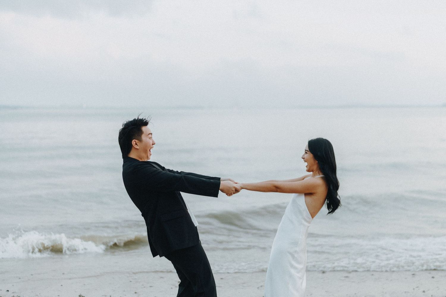 Romantic couple dancing joyfully on a misty beach during sunset with waves crashing behind them.