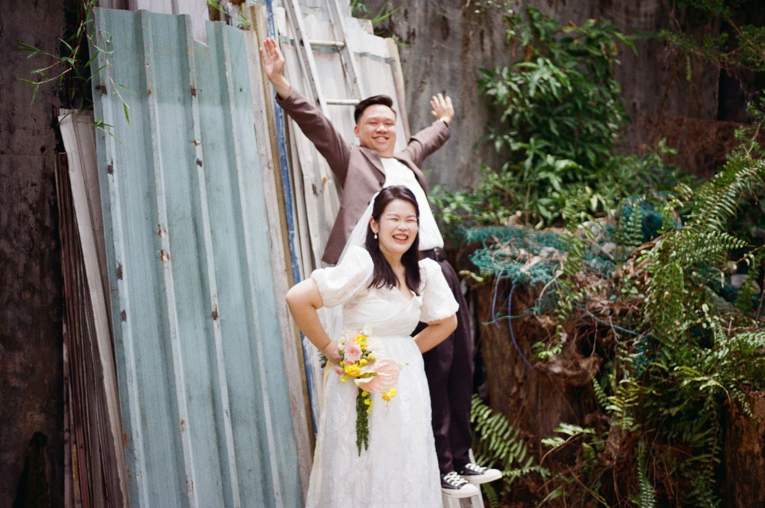 A joyful couple poses together outdoors against a rustic fence, with the bride holding a yellow bouquet in her white dress.