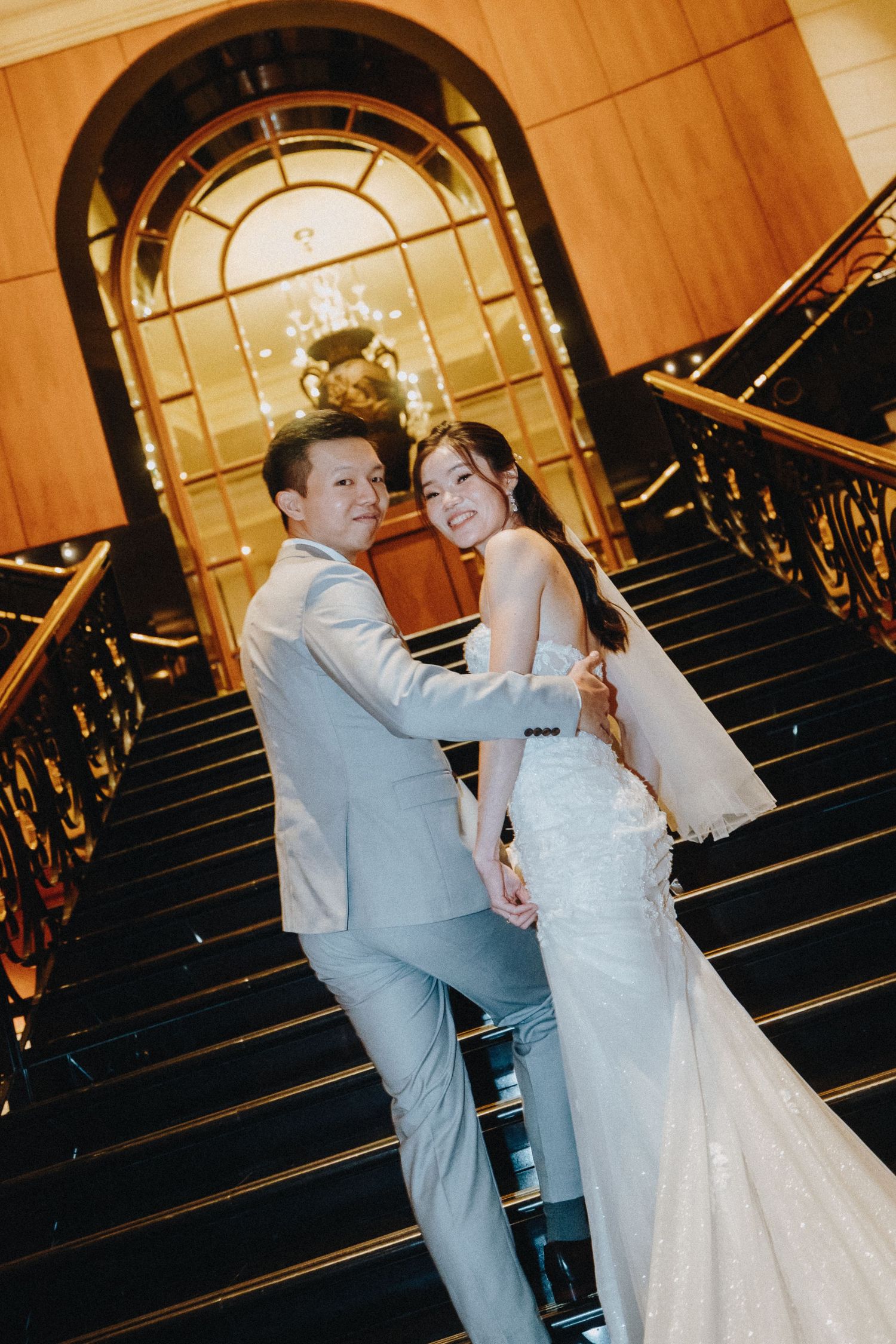 A wedding couple poses romantically on an elegant staircase with an arched window and golden lighting.