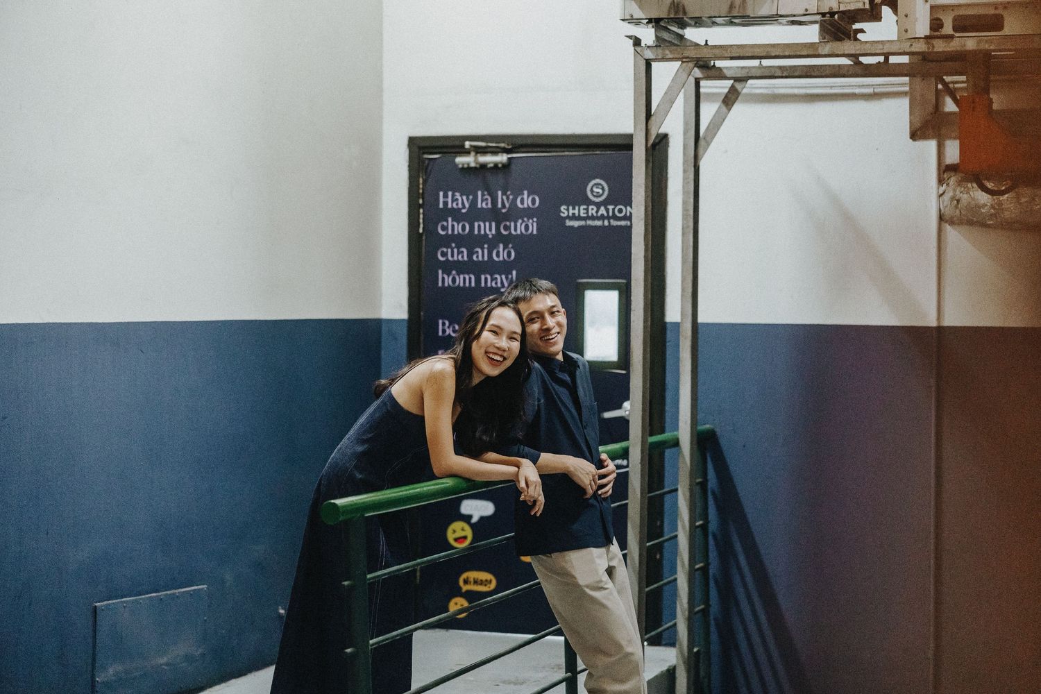 A couple sits together on a green bench in a hallway with blue and white painted walls and industrial decor.