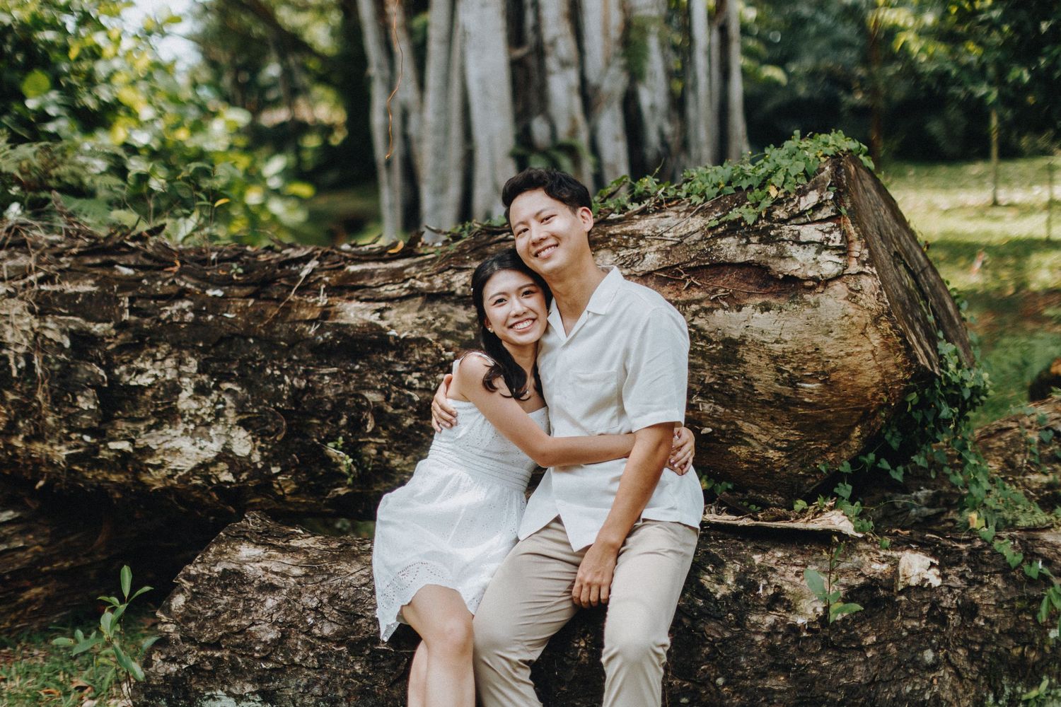 Couple sitting together on fallen log in wooded area during outdoor photoshoot.