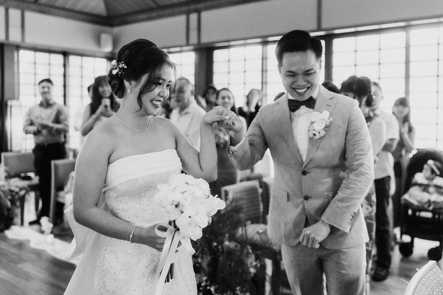 Wedding ceremony attendees gather in bright indoor venue with large windows.