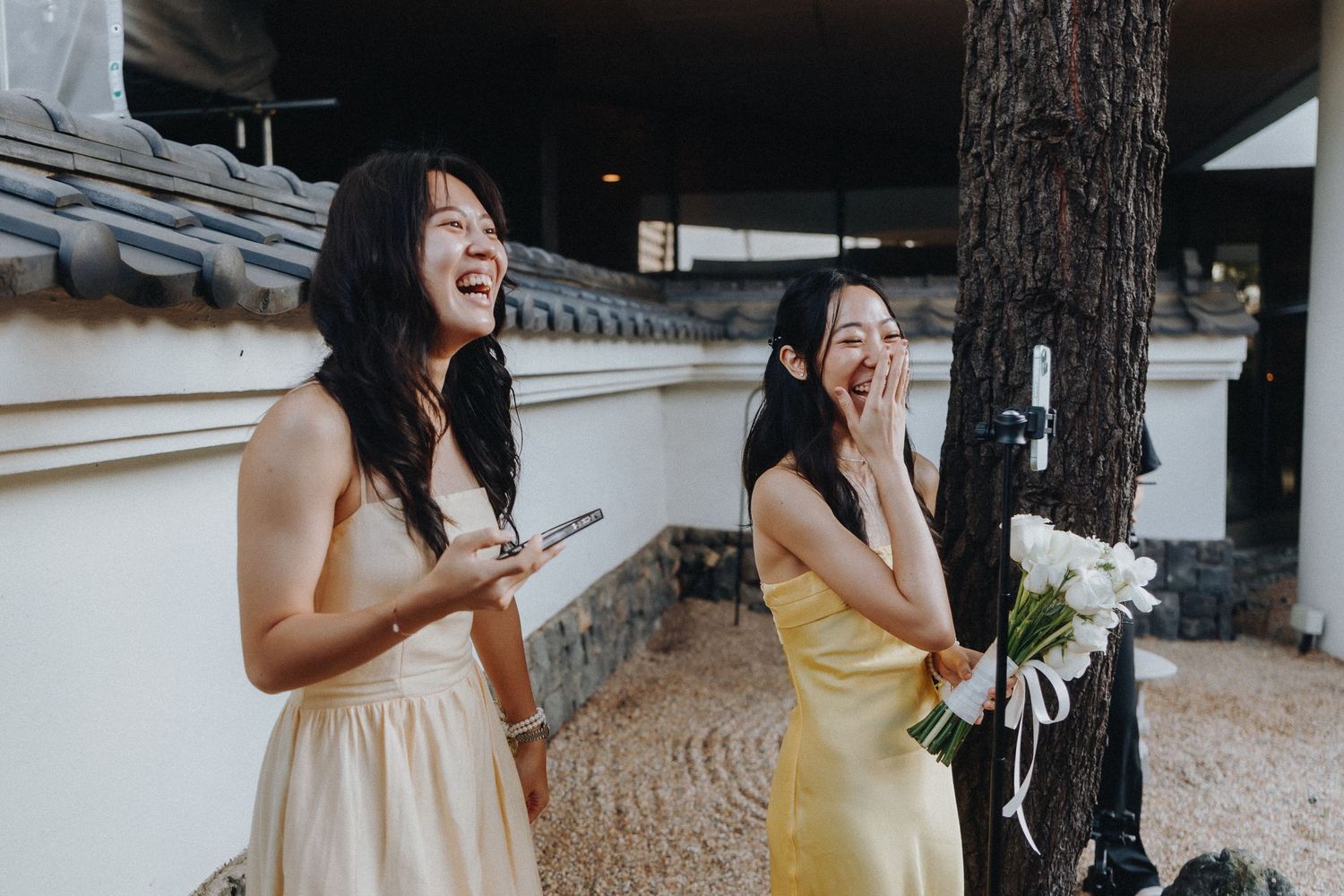 Two bridesmaids in formal attire share a joyful laughing moment at an outdoor wedding venue.