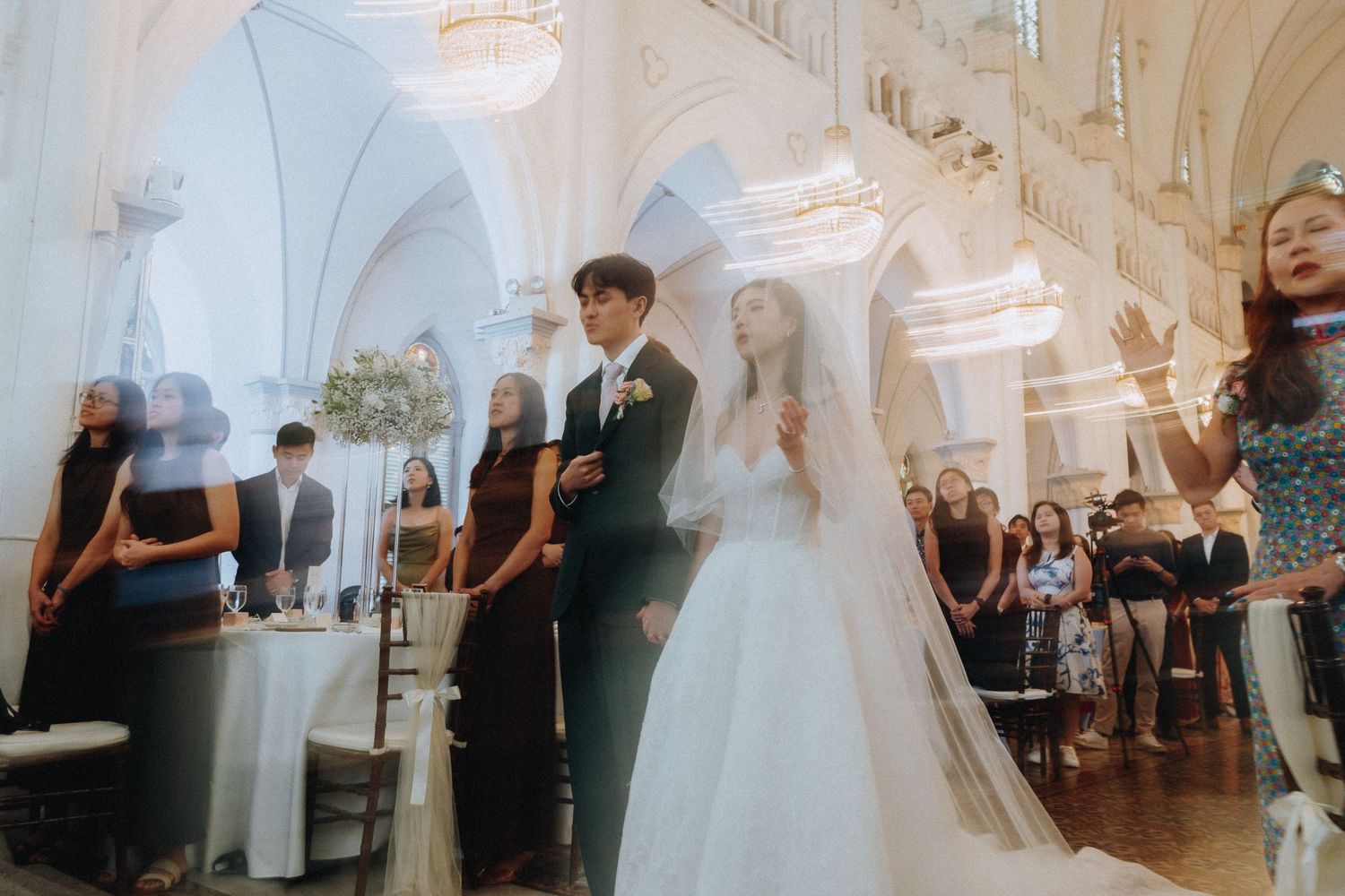 Bride and groom walk down the aisle in an elegant white church surrounded by guests during a wedding ceremony.