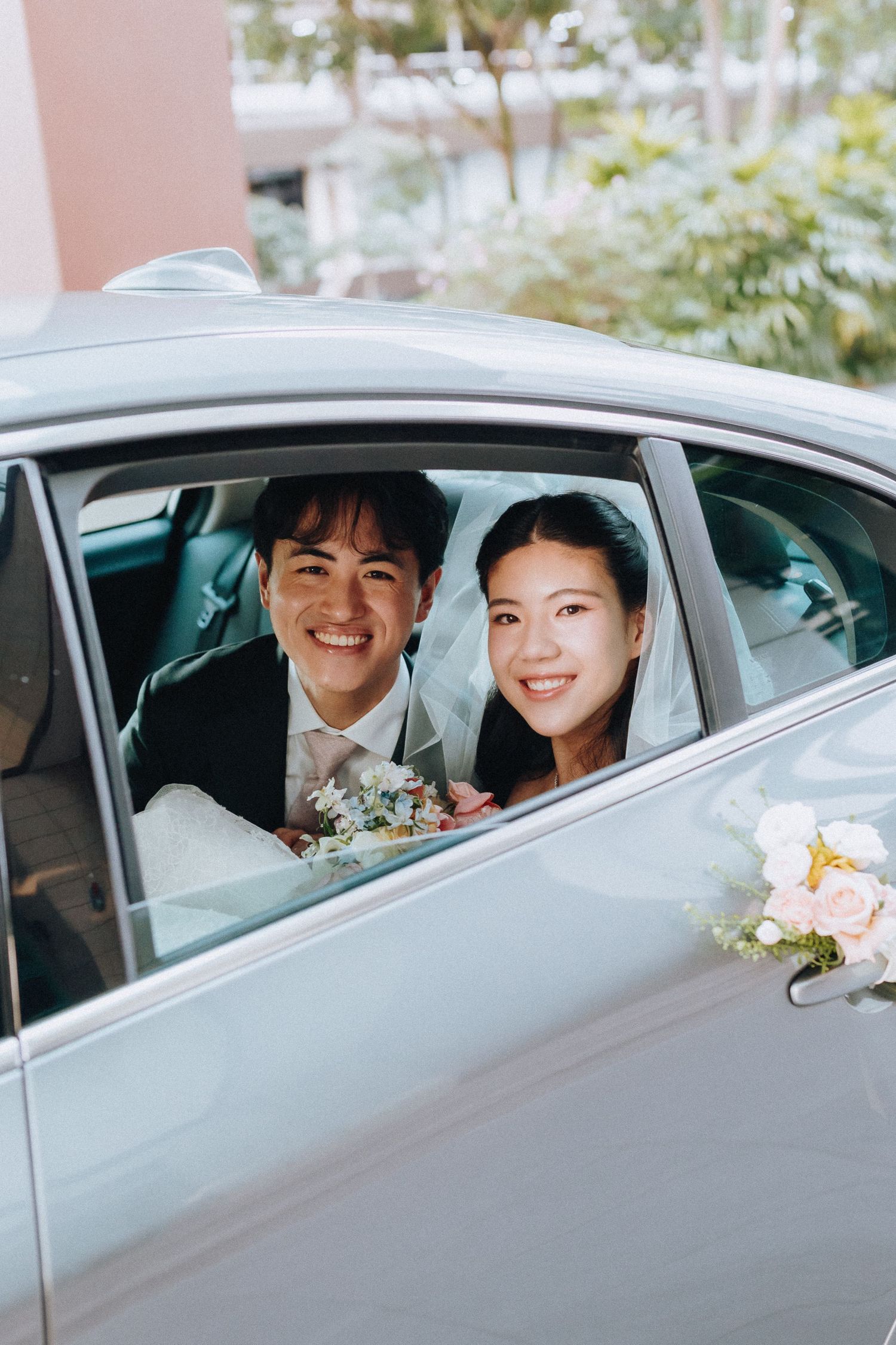 Smiling Asian bride and groom sitting in a decorated wedding car, bride holding a floral bouquet.