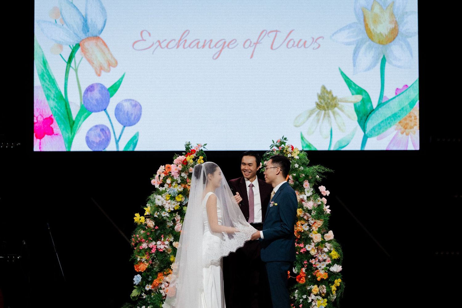 Couple exchanging vows at floral arch altar beneath Exchange of Vows screen at wedding ceremony.