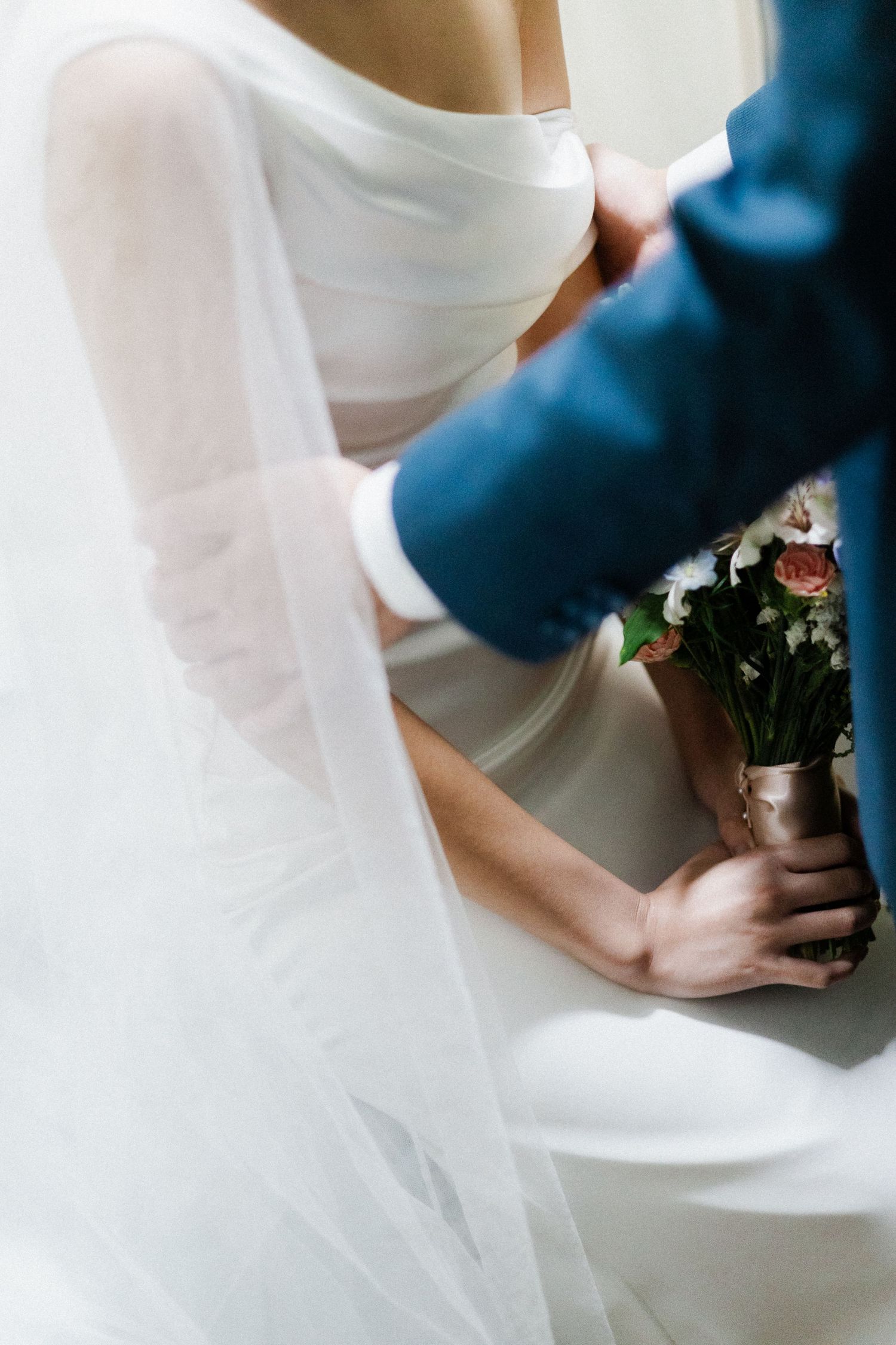 Groom in blue suit holding bride's bouquet of flowers beside her white wedding gown and veil.