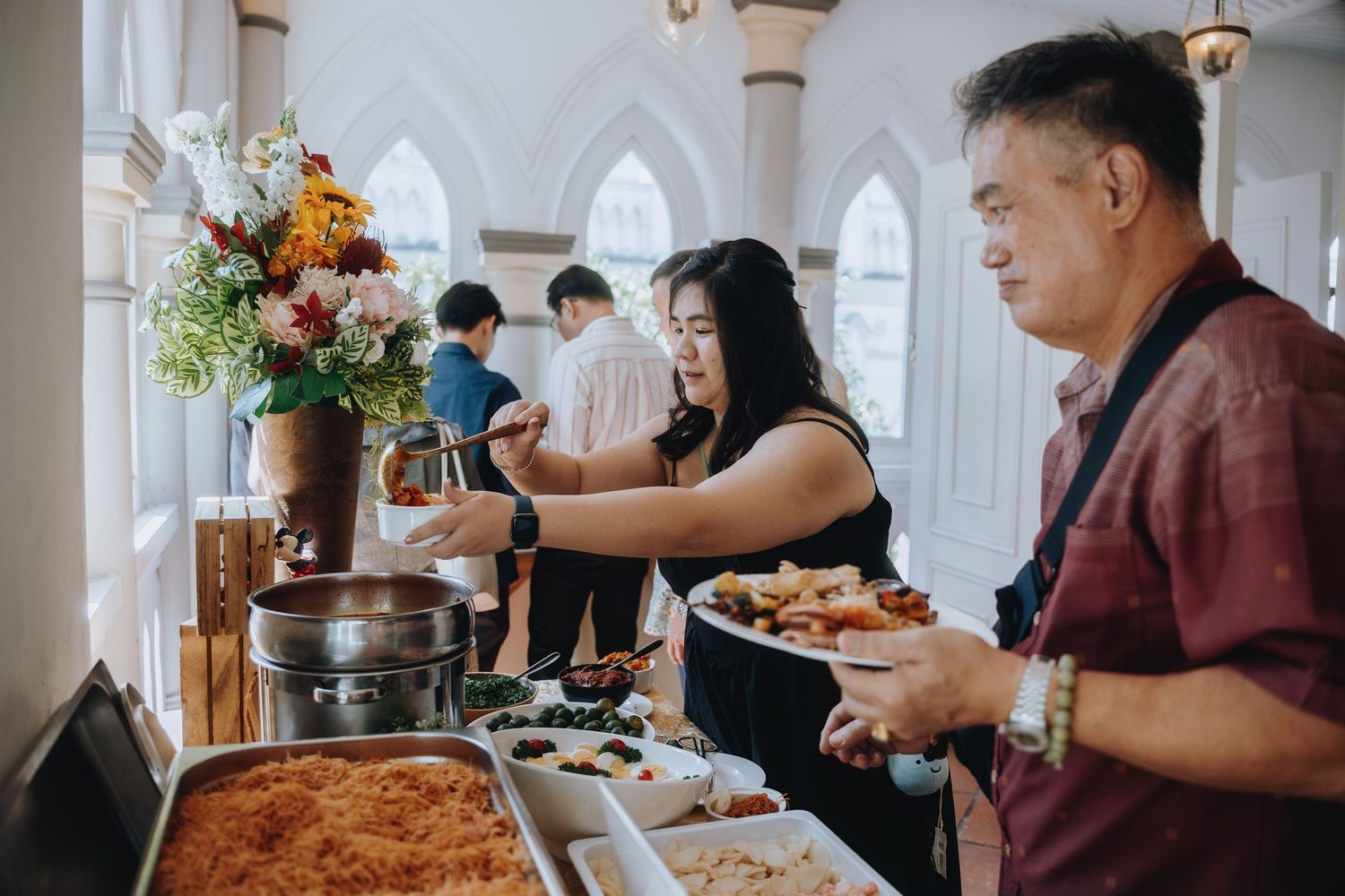 Guests serve themselves at an elegant wedding reception buffet with colorful dishes and floral arrangements.