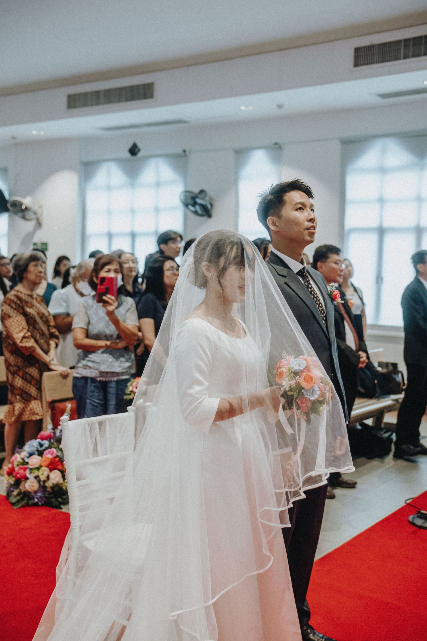 Bride in white veil and groom in suit walk down red aisle at church wedding.