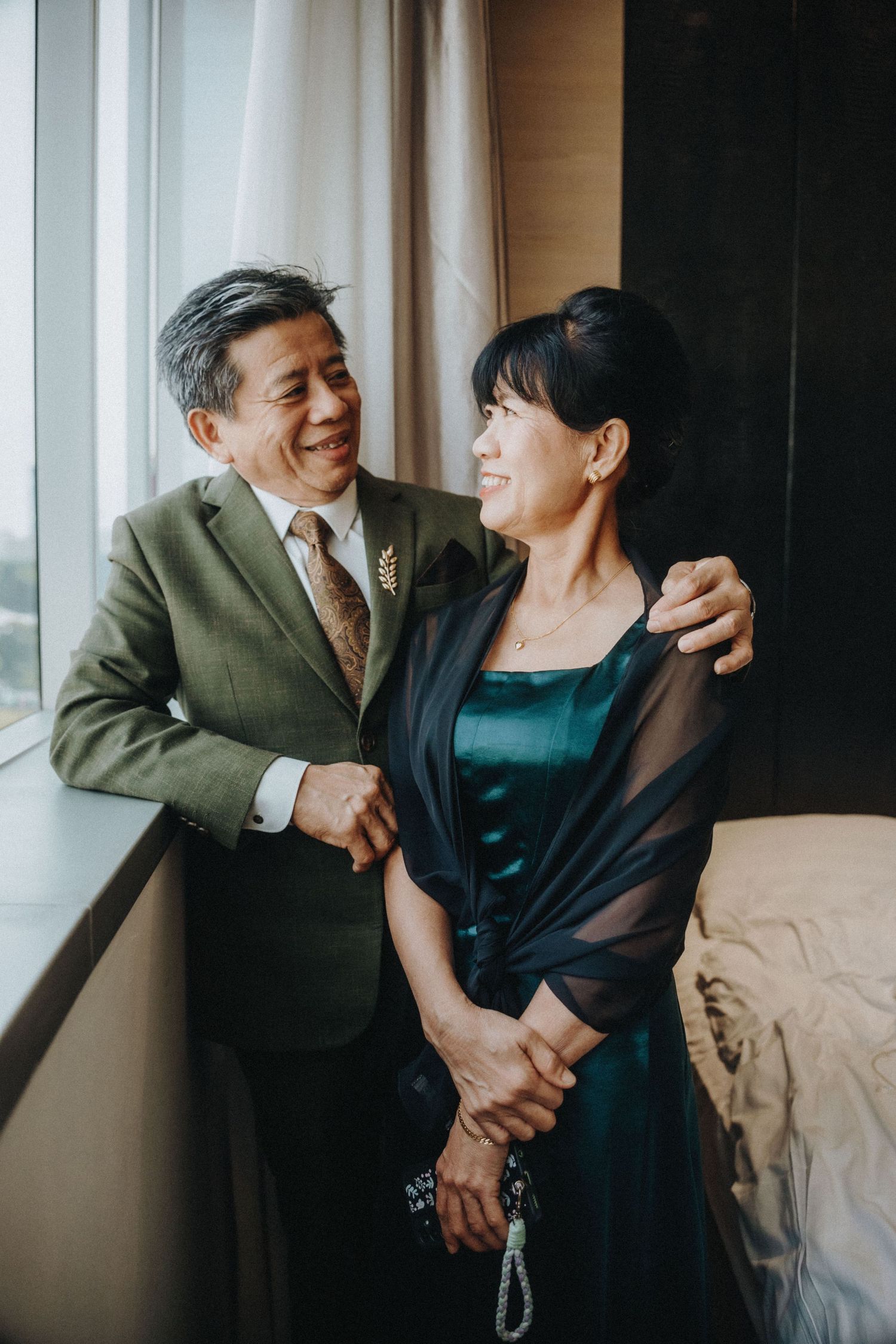 Elegant older Asian couple smiling together by a window, man in green suit with arm around woman in teal dress.