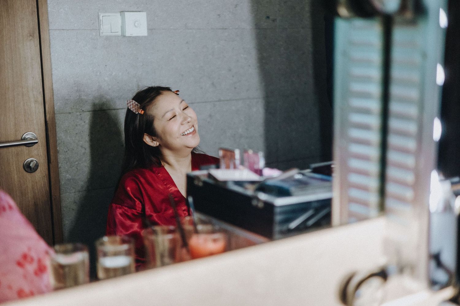 A joyful young woman in red satin laughs at a vanity mirror surrounded by makeup and candles.