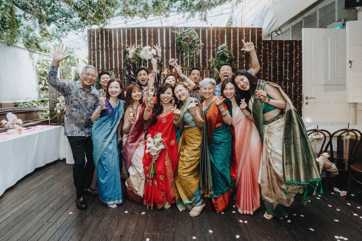 Large group of wedding guests in vibrant sarees and batik shirts posing joyfully outdoors.