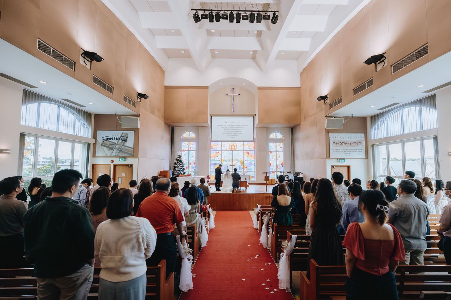 Guests stand during a church wedding ceremony with red carpet aisle and stained glass windows.