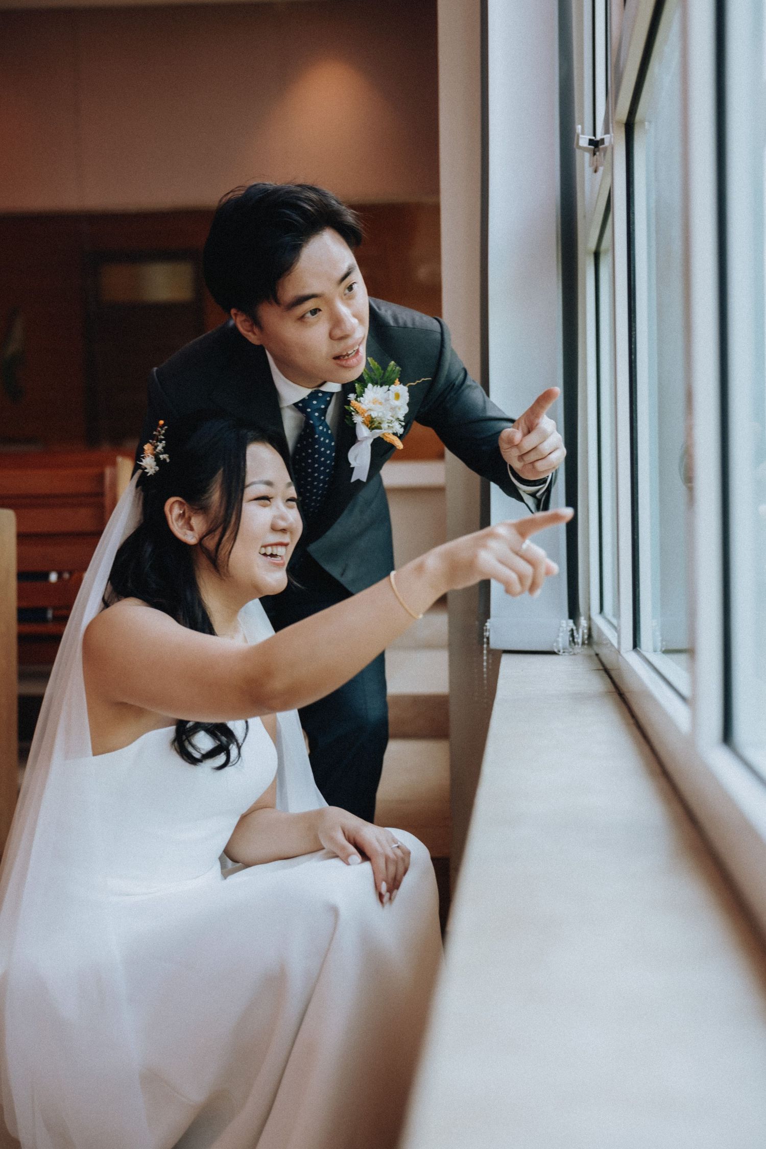 A smiling bride and groom in wedding attire point out a window together during their wedding day.