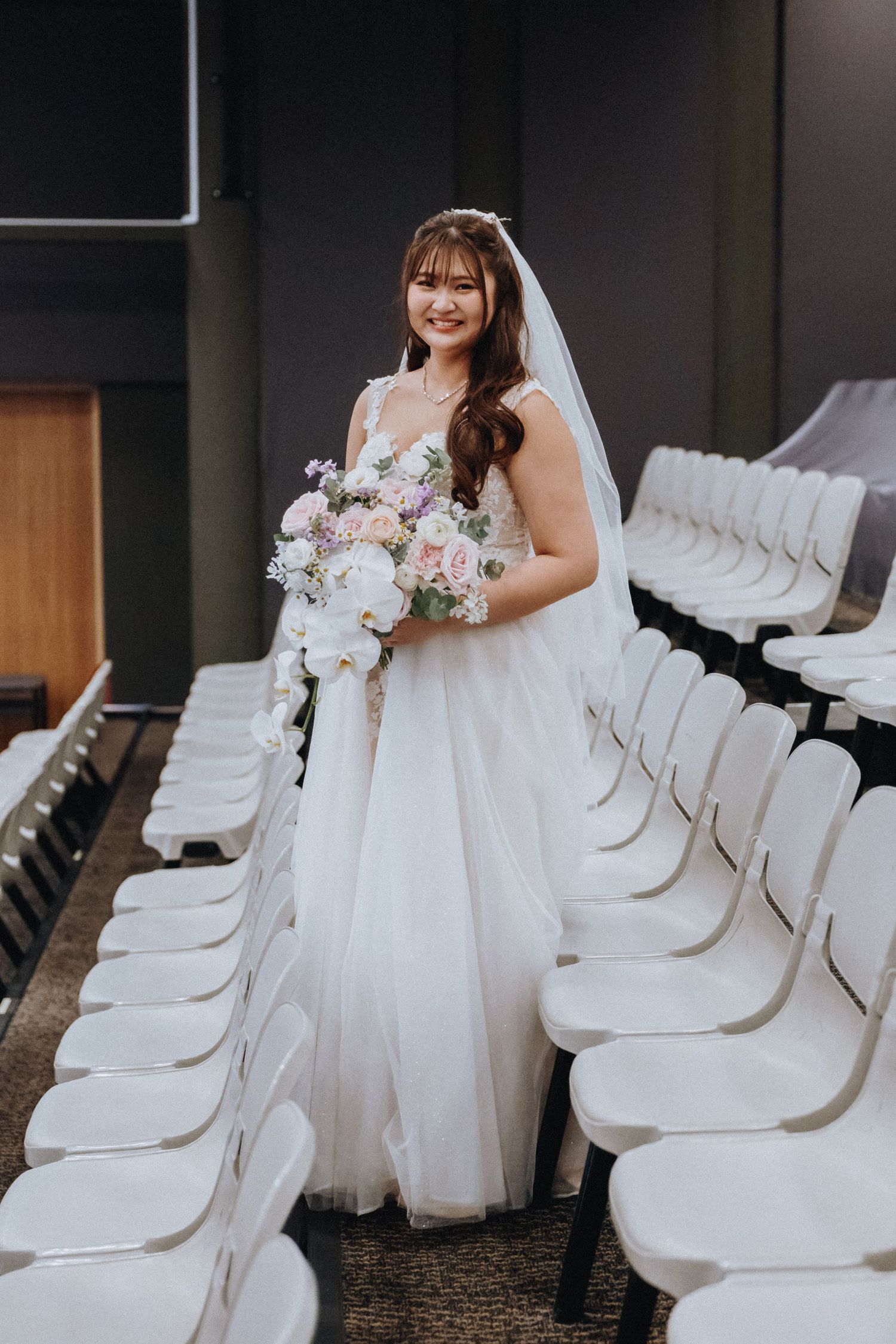 A bride wearing a white tulle gown holds a pink and white bouquet while standing in an auditorium.