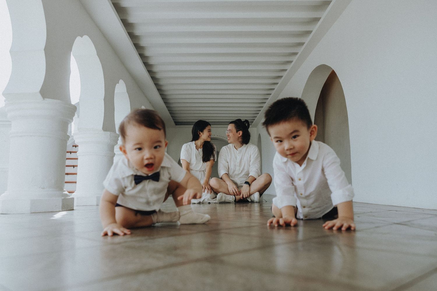 Children crawl playfully on floor while parents sit in background of white arched hallway.