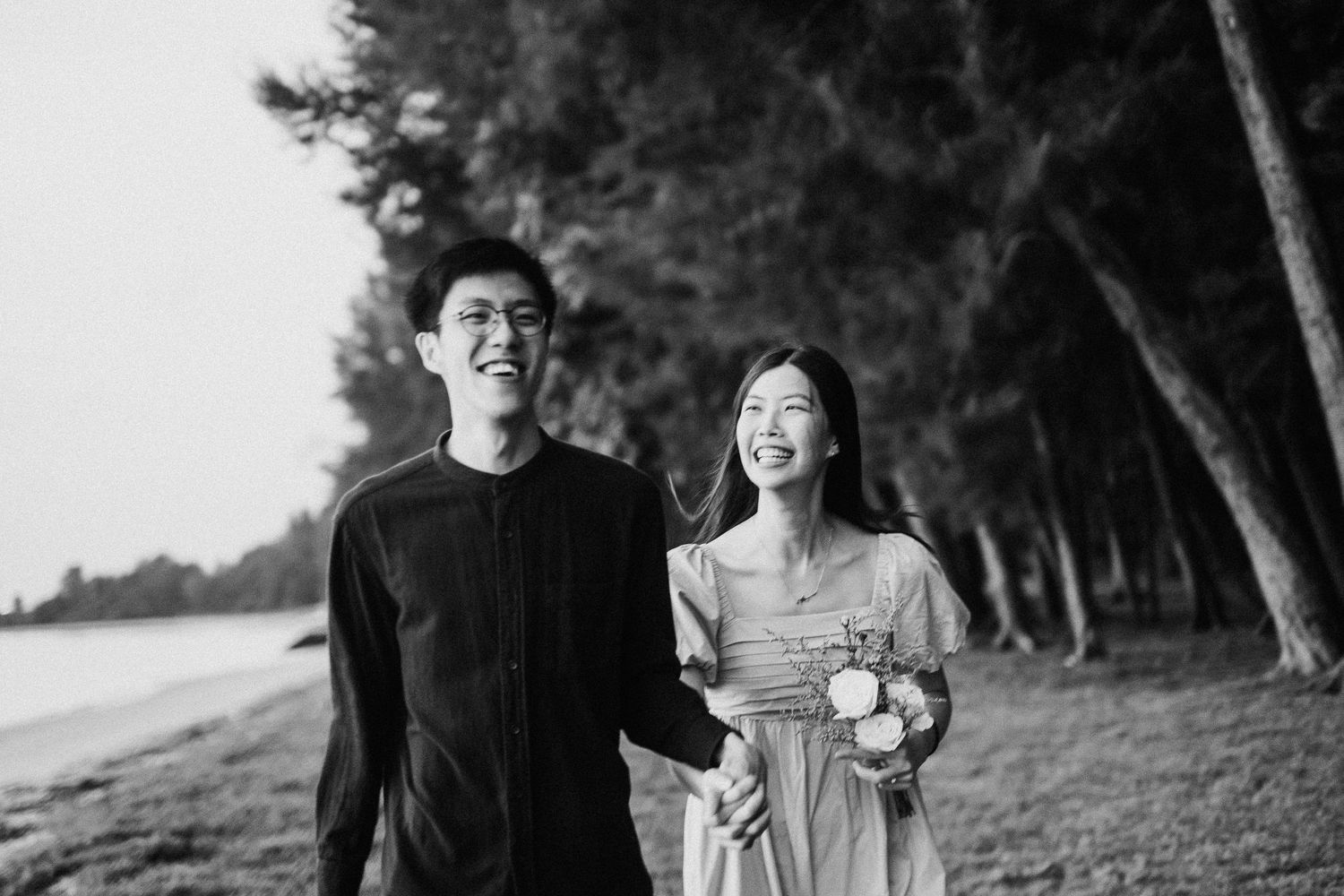 A couple walks hand in hand through a serene wooded path captured in black and white.