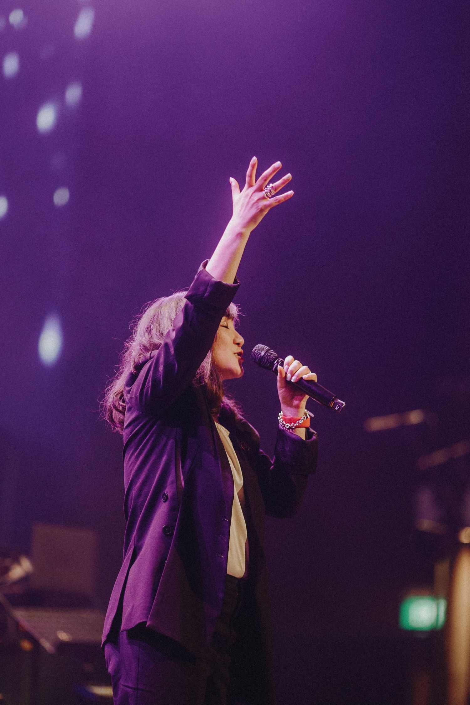 Performer in purple blazer sings passionately on stage with dramatic lighting during live concert performance.