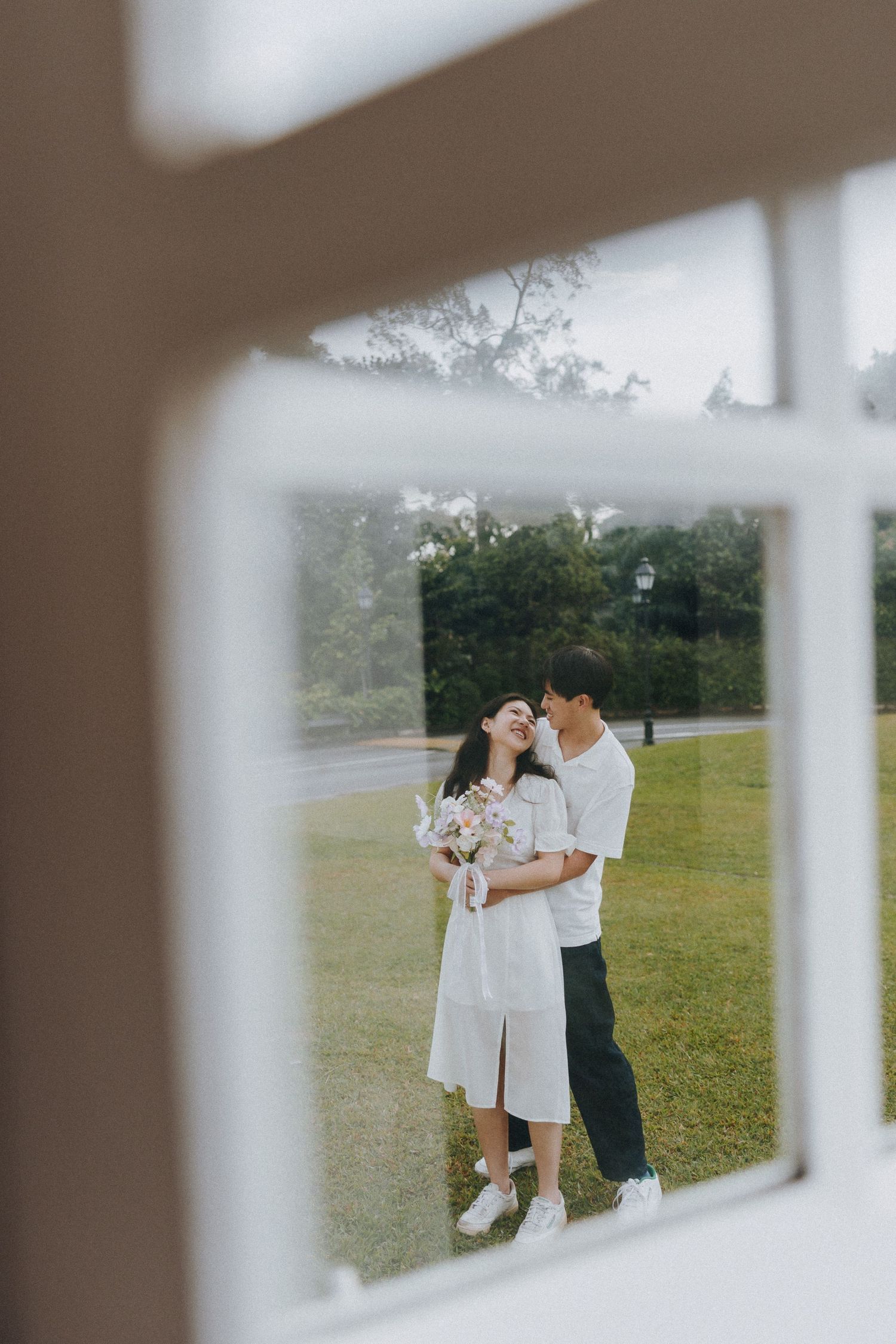 A couple in white outfits viewed through a window frame on a sunny lawn.