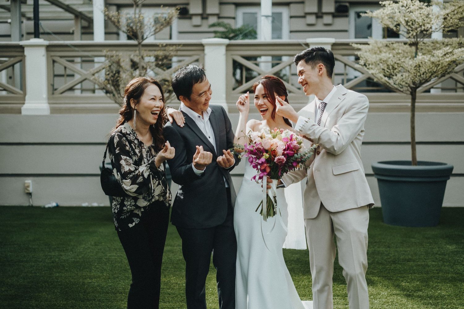 Wedding party shares joyful laughter together during outdoor garden celebration by a historic building.