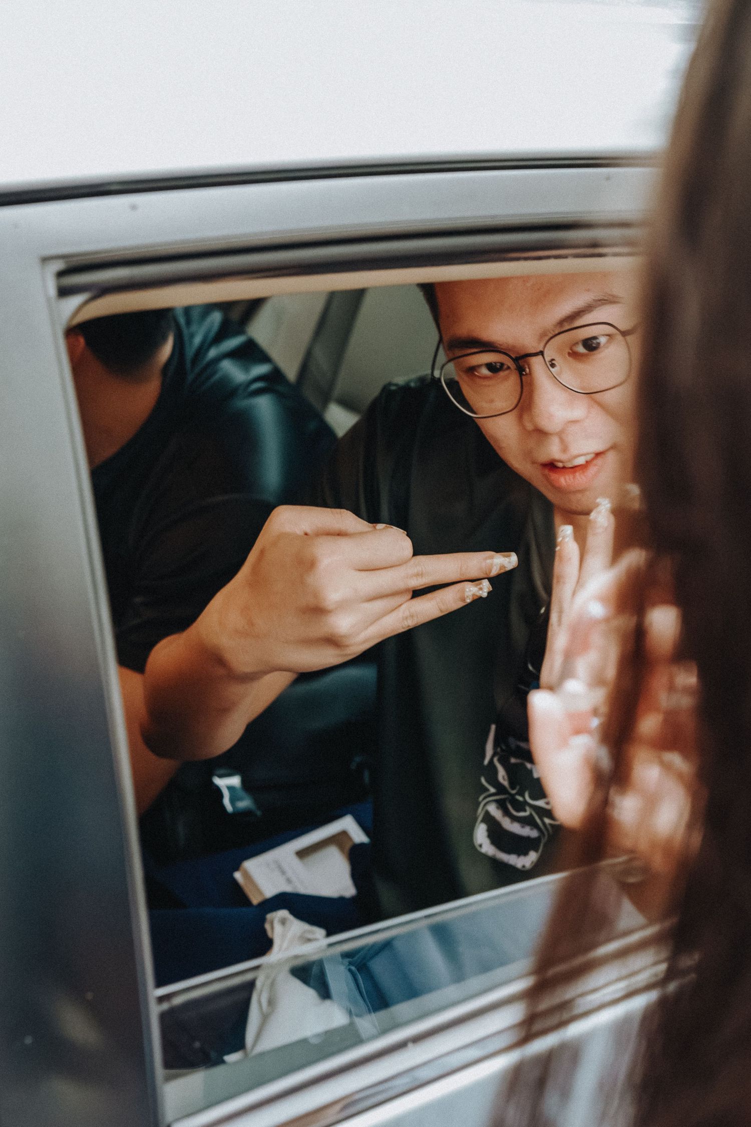 Through a car window, someone wearing glasses is seen adjusting their hair.