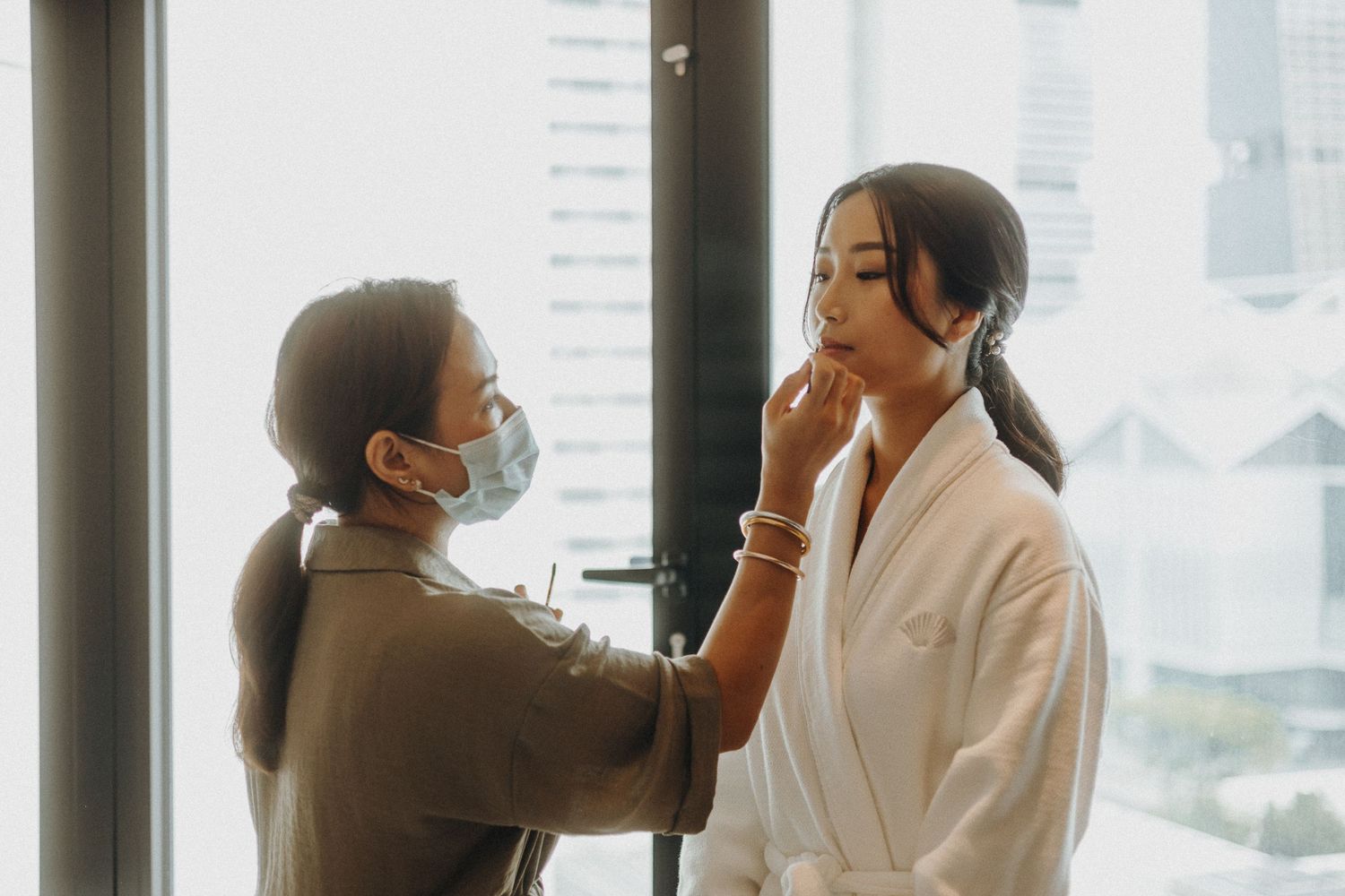 Wedding preparations in a bright hotel room with bridesmaids helping with white gowns and robes.