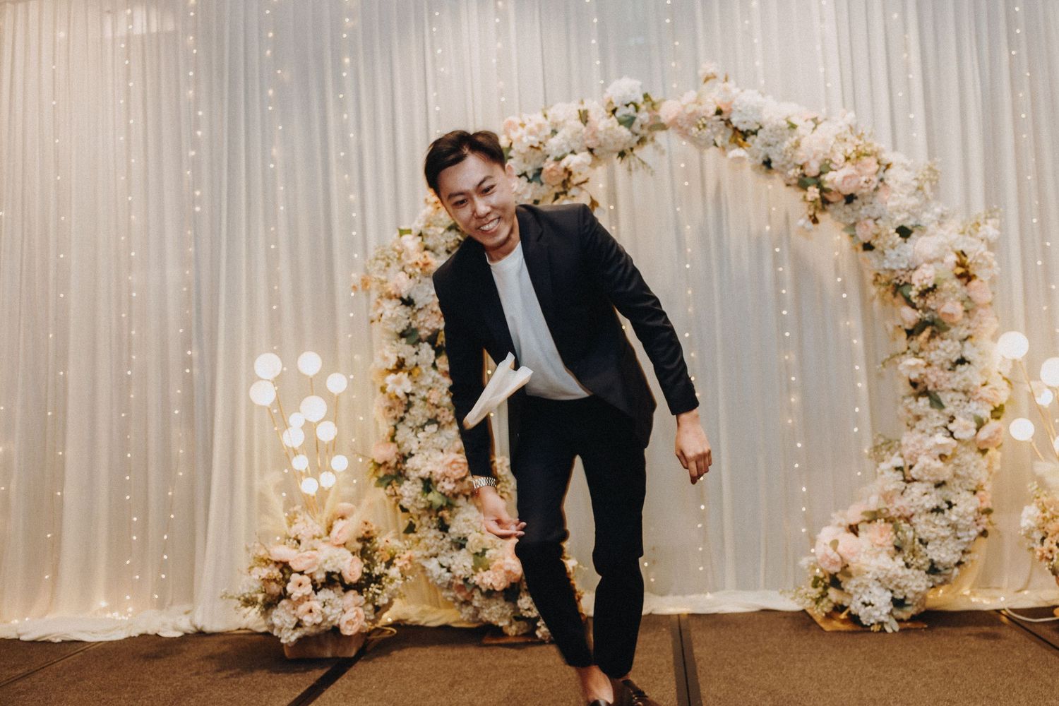 A person in a black suit strikes a playful pose under a floral wedding arch.