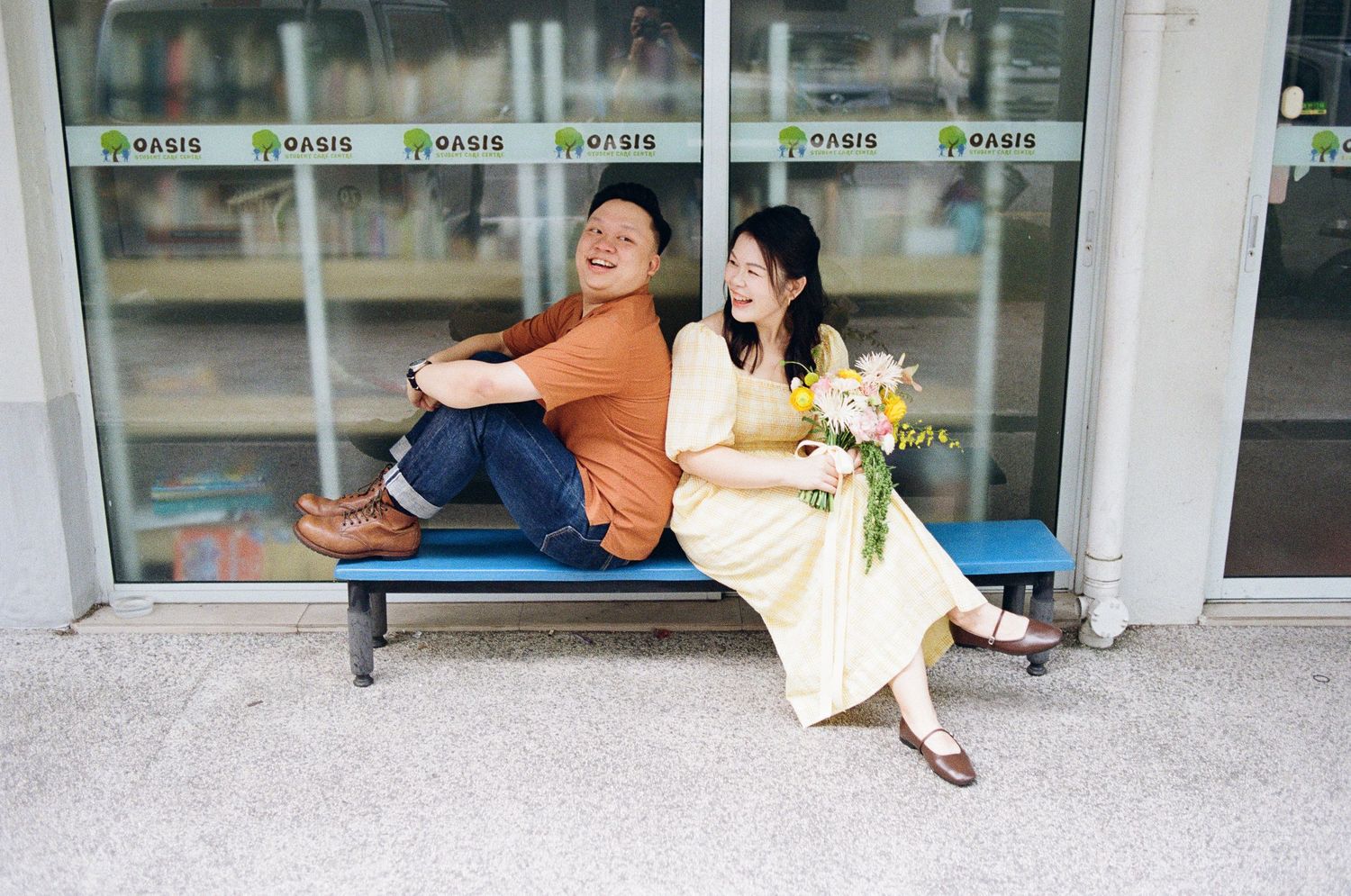 A couple in casual attire sits back-to-back on a bench outside a storefront.