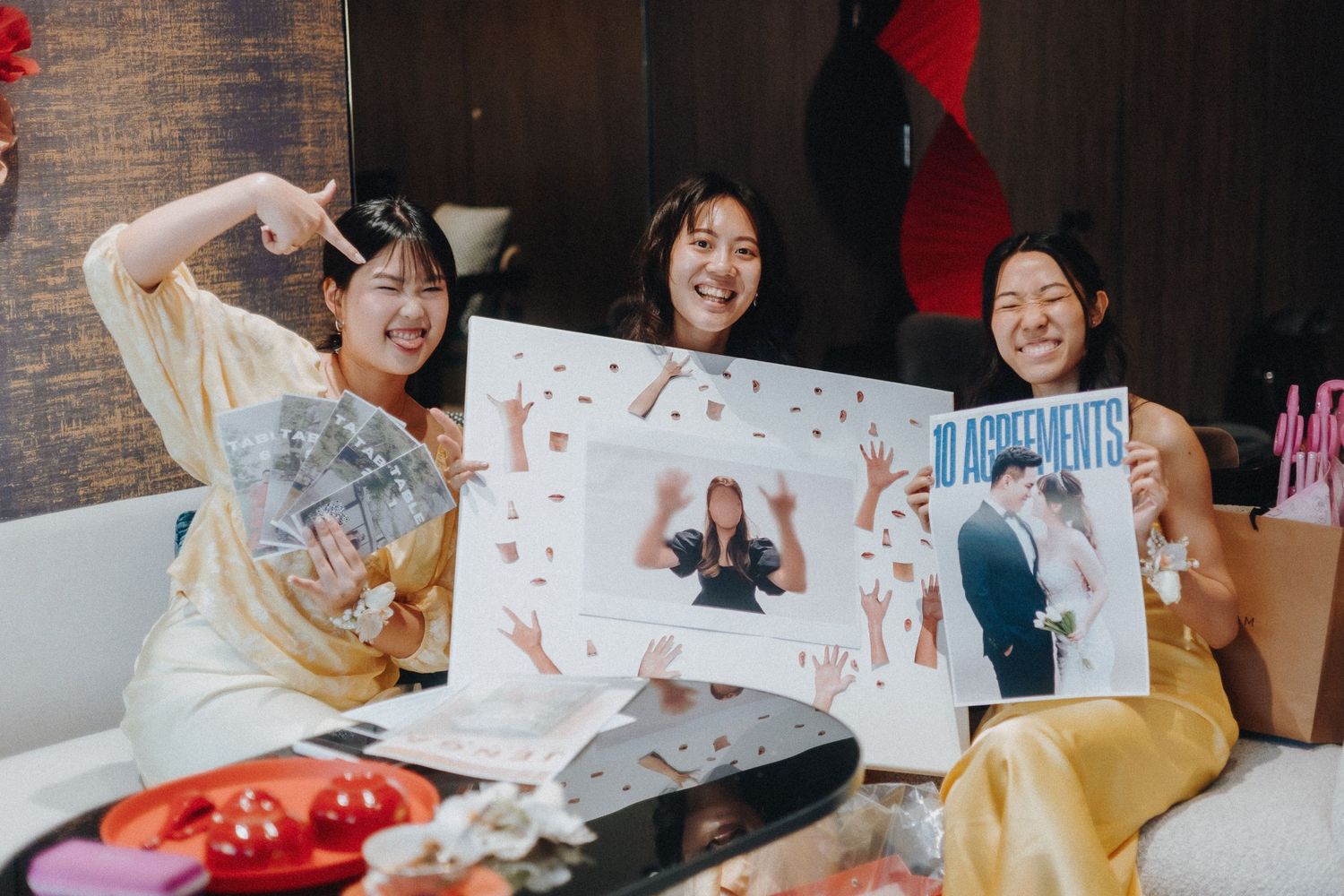 Three friends smile while showing photo prints and artwork while sitting on a couch together.