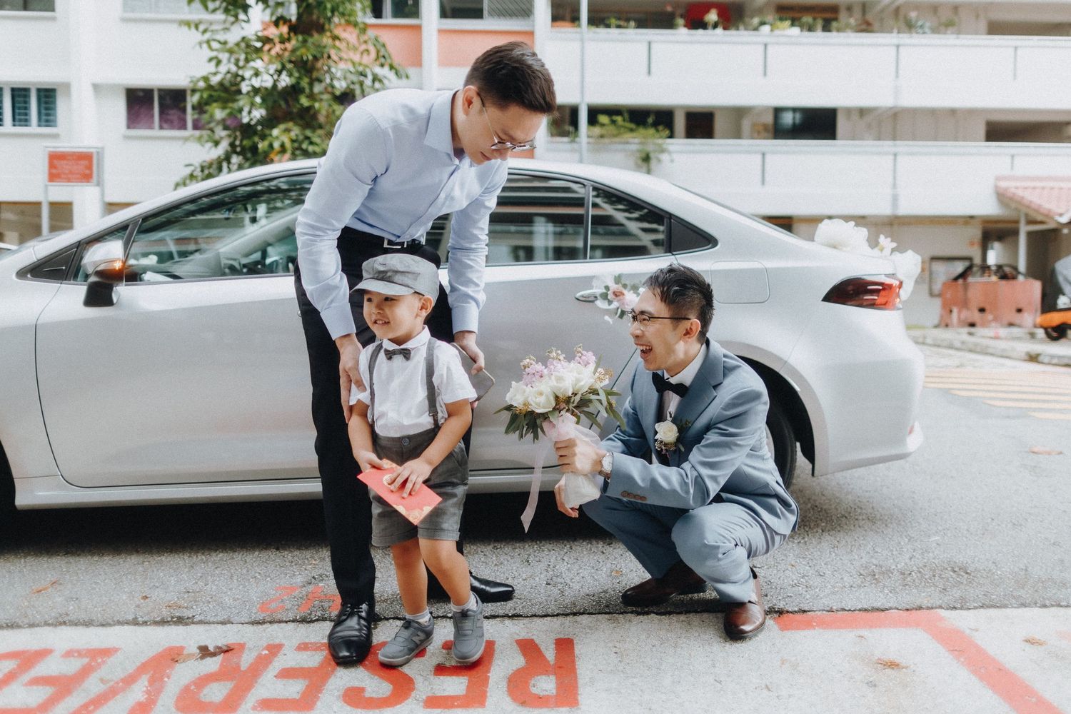 Three people gather around a silver car parked on the street during a casual outdoor moment.