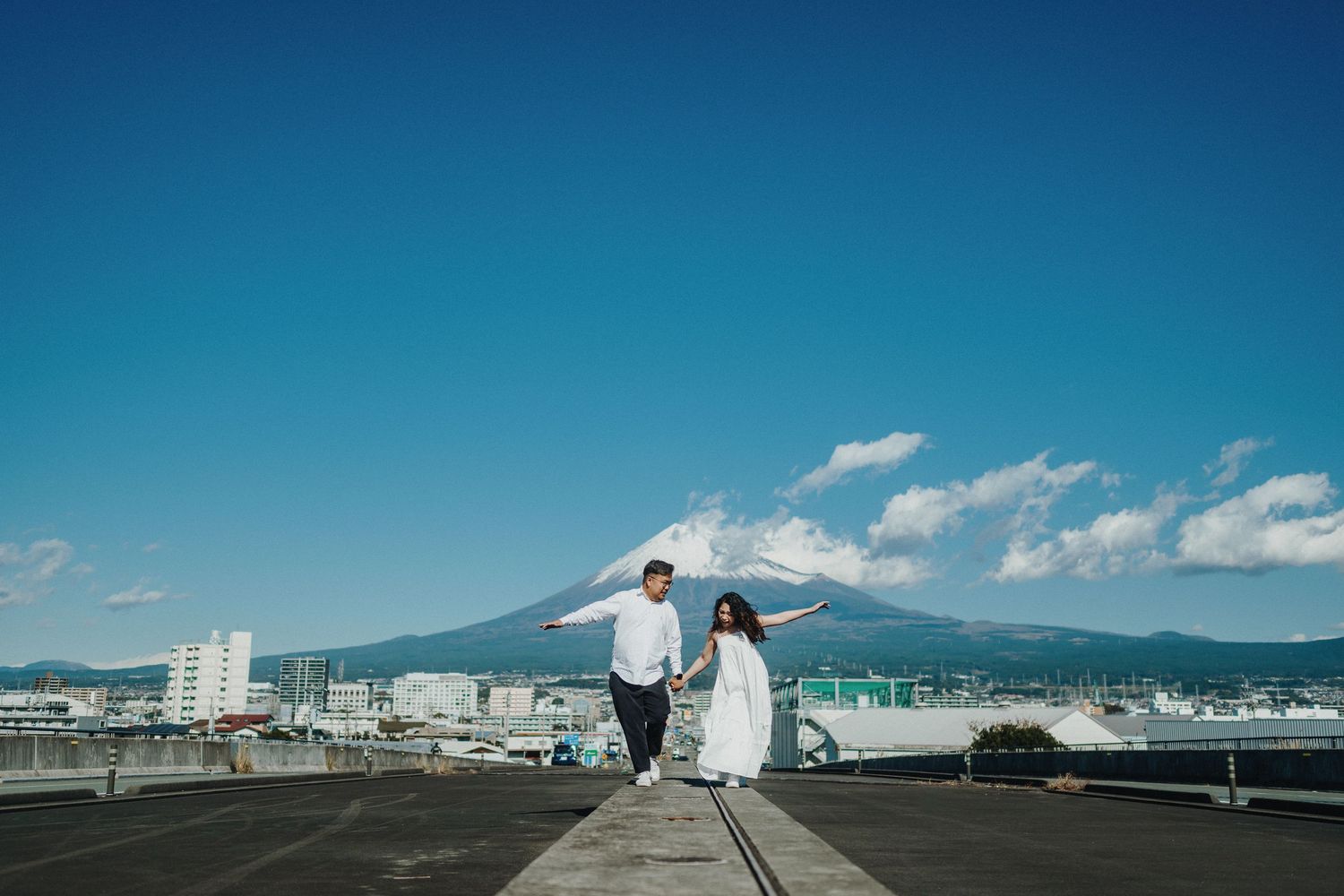 A couple in wedding attire stands on a pier with Mount Fuji and blue skies in the background.